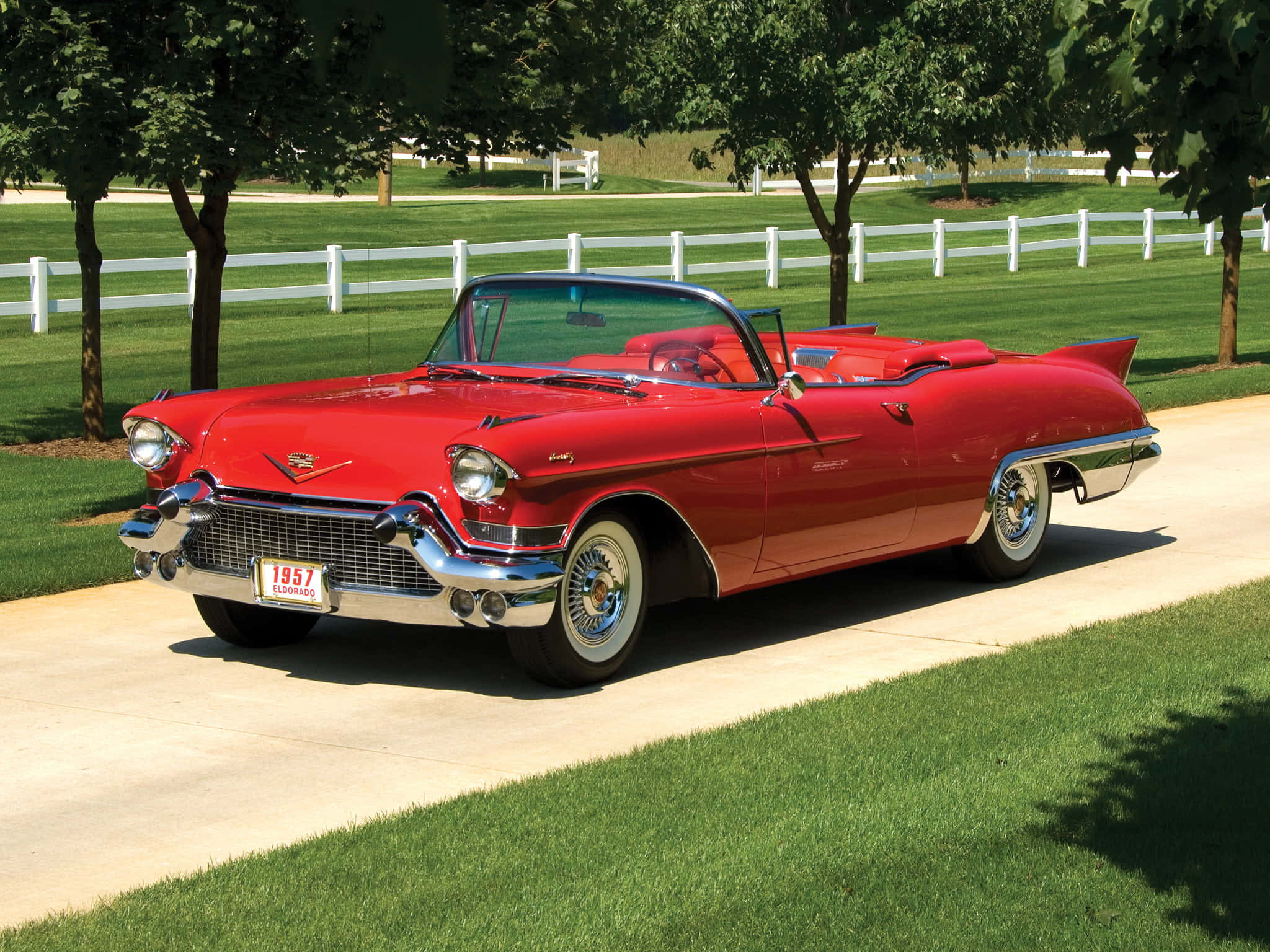 Cadillac Eldorado 桌布 壁紙 — 一架光澤的紅色1957年凱迪拉克敞篷車停泊在鋪砌的小路上，旁邊是茂密的綠草和白色圍欄，在明亮的陽光下。