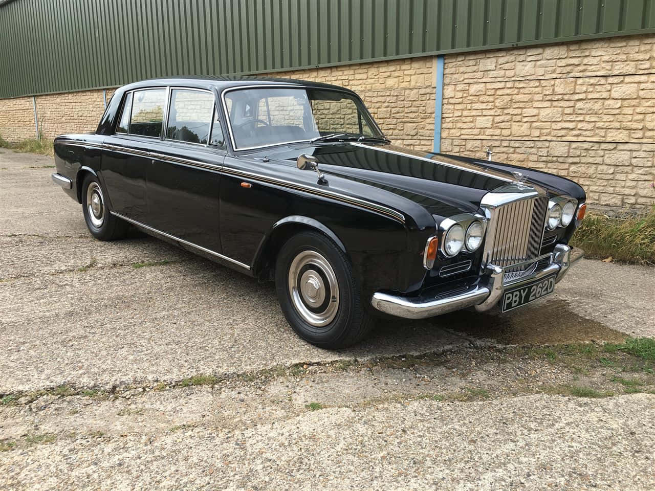Bentley T1 桌布 壁紙 — 一輛光澤黑色經典勞斯萊斯在明亮的日光下，停放在碎石地上，旁邊是石頭和金屬牆。