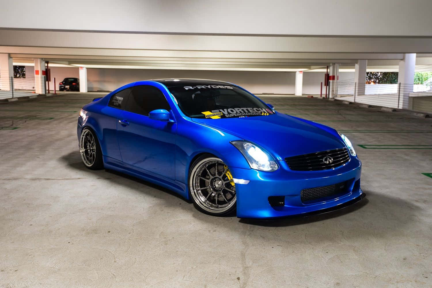 Infiniti G35 桌布 壁紙 — 一輛光澤藍色改裝跑車，配備定制輪圈和貼紙，停放在混凝土停車場的人工照明下。