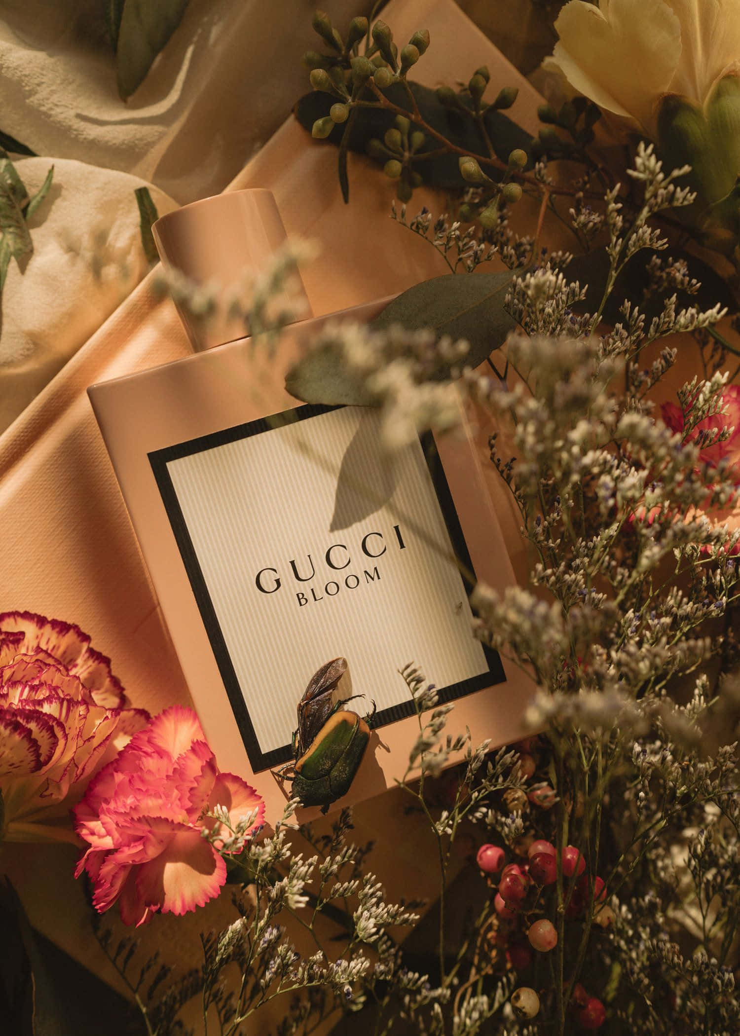 「 effortlessly elegant and captivating - a fragrance to make you unforgettable.」 壁紙 — Gucci Bloom 香水瓶置於柔軟布料和鮮豔花朵中，沐浴在溫暖的金色光線下。