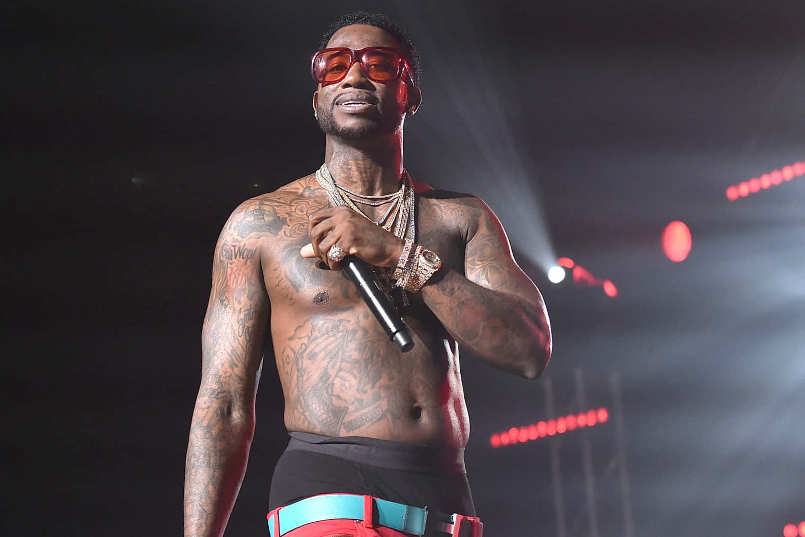 Gucci Mane 舞台演出 壁紙 — 一位赤裸上身、有紋身的說唱歌手，戴著紅色太陽眼鏡，在舞台燈光下拿著麥克風。