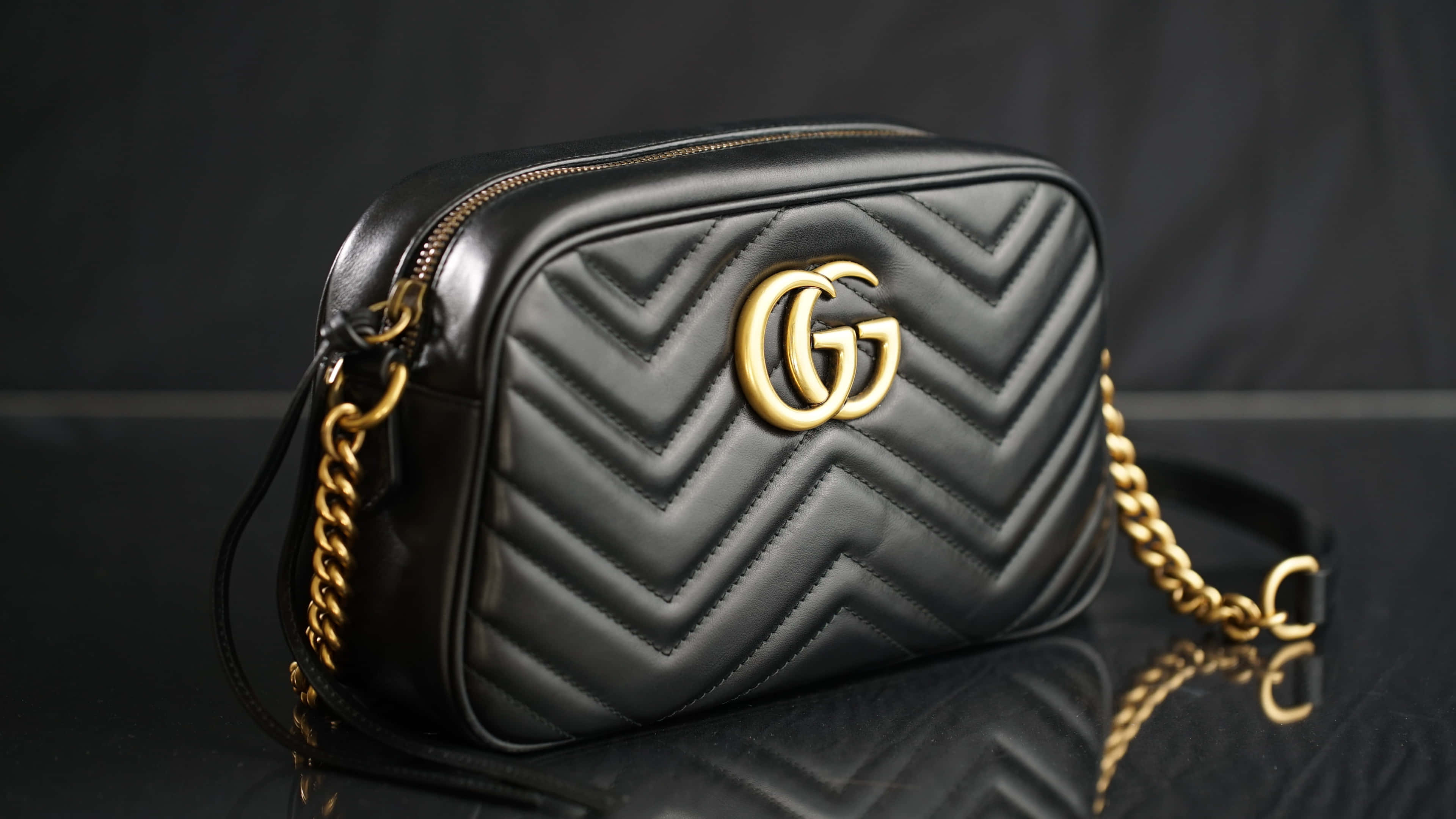 Gucci黑色手袋4K 壁紙 — 一個黑色絎縫Gucci手袋，配金色鏈條肩帶，放在深色背景的反射表面上。