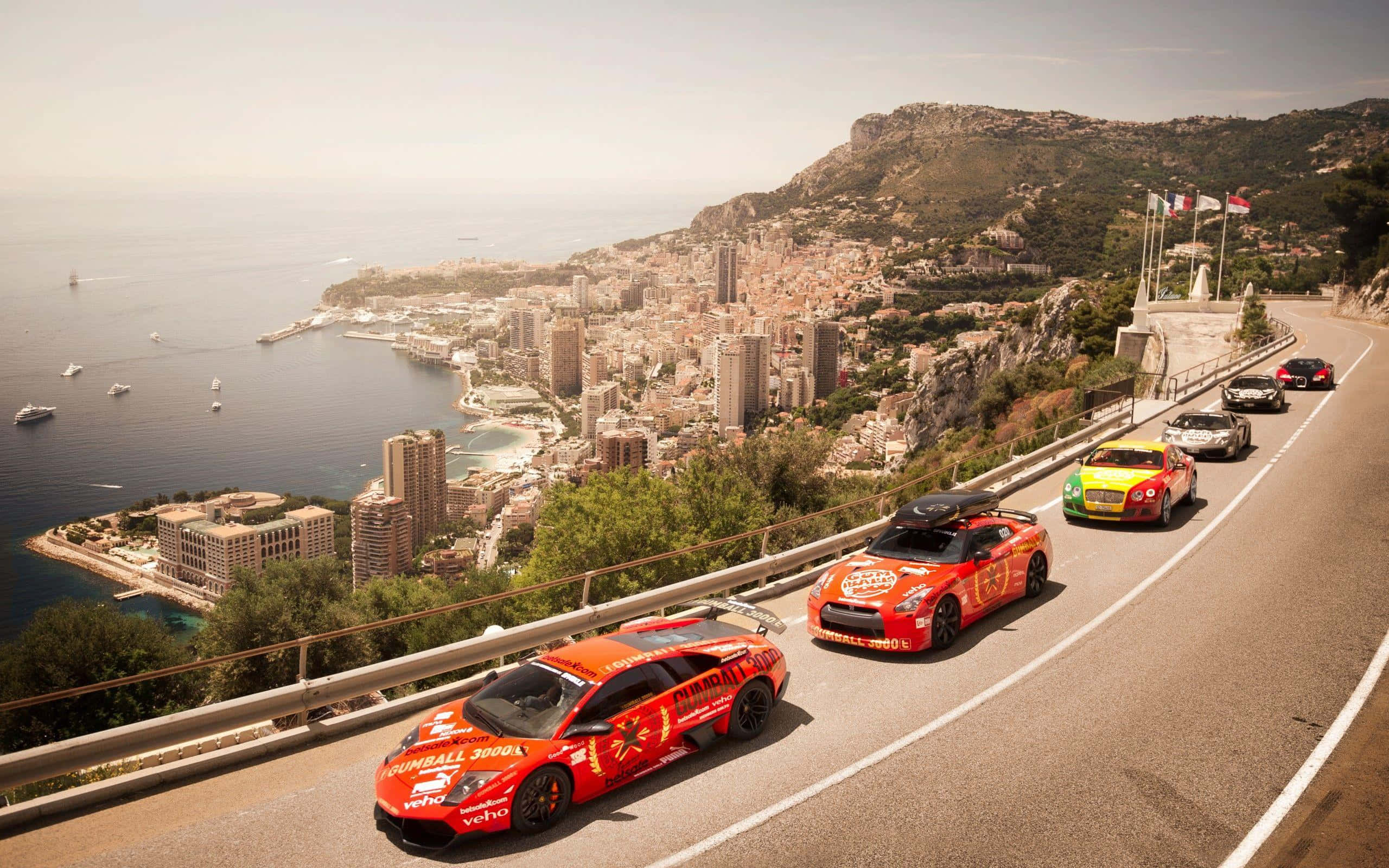 Gumball 3000 摩納哥賽車活動 壁紙 — 色彩繽紛的賽車沿著海岸山路飛馳，俯瞰摩納哥密集的城市和閃閃發光的藍色大海。