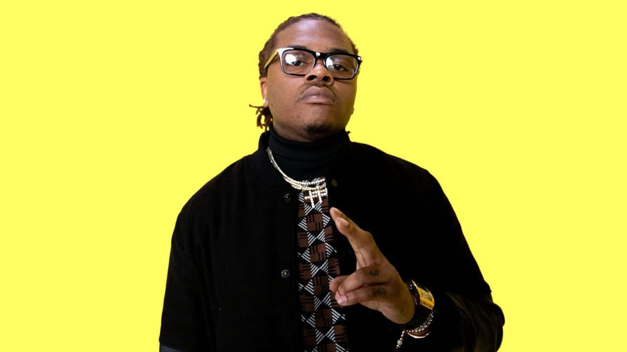 Gunna 創意訪問攝影 壁紙 — 一名戴眼鏡、打著圖案領帶的男子，在明亮的黃色背景前自信地做手勢。