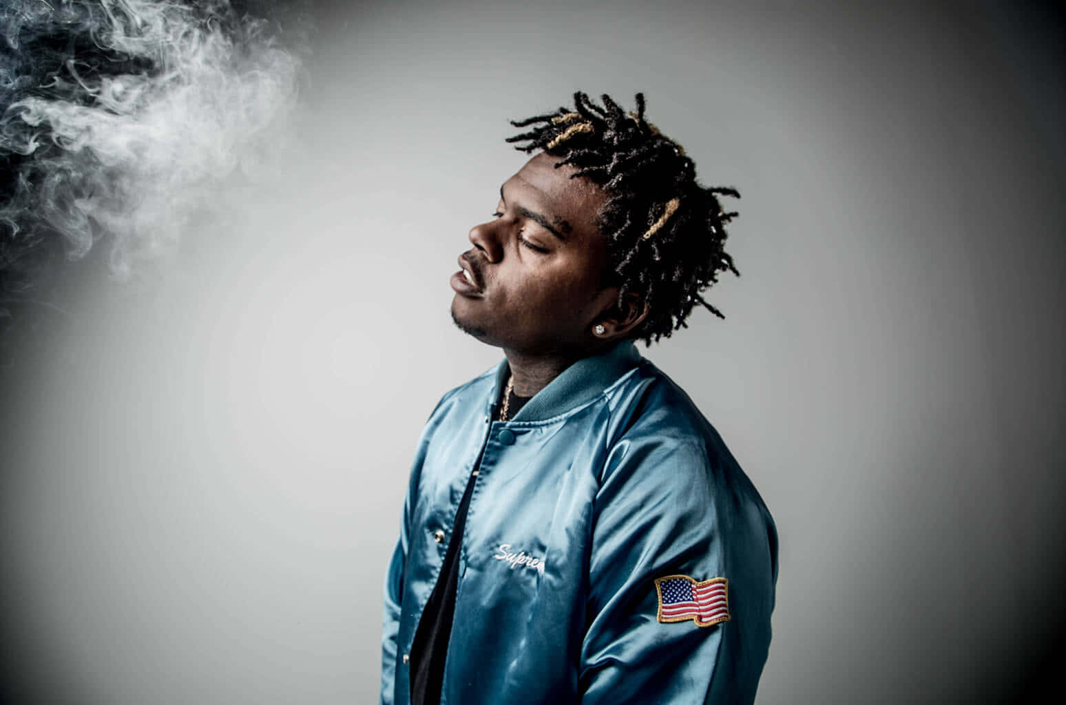 Gunna 美國說唱天王 壁紙 — 一名身穿藍色緞面外套的男子在灰色攝影棚背景前，在戲劇性燈光下呼出煙霧。
