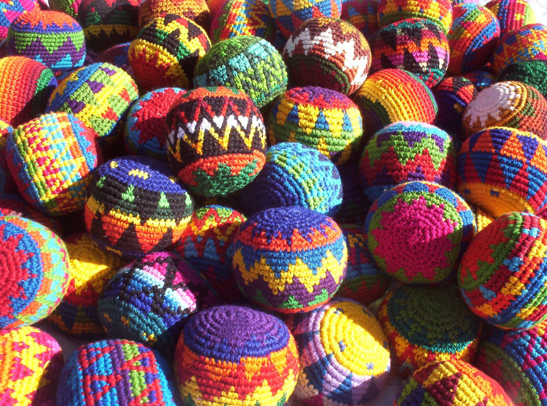 織物螺旋Hacky Sack，展現技巧與文化 壁紙 — 鮮豔的手工鉤針球體，帶有鮮明的幾何圖案，在明亮的陽光下堆疊在一起。
