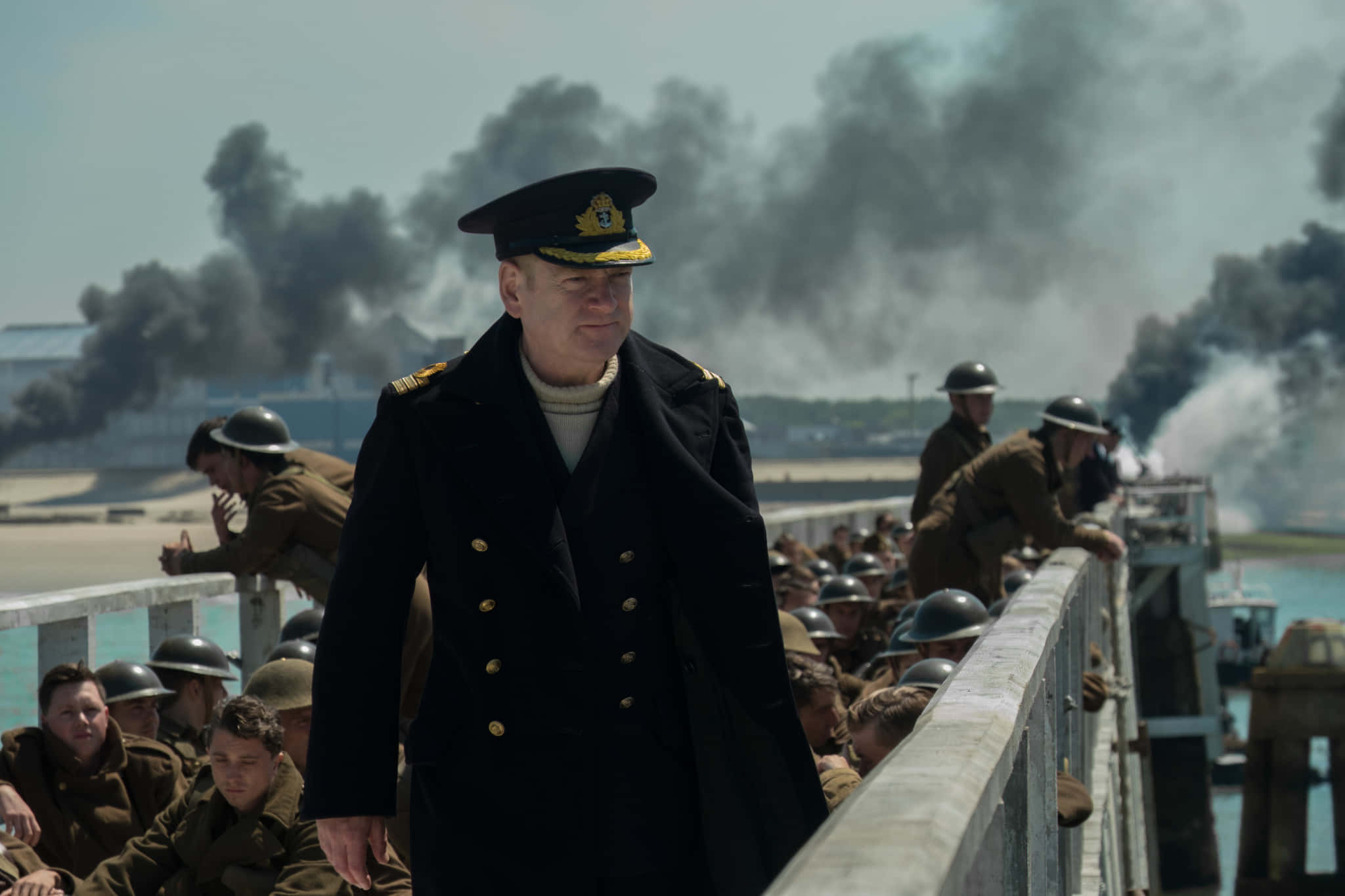 Dunkirk 桌布 壁紙 — 一名身穿黑色外套的海軍軍官莊嚴地走在碼頭上，戴頭盔的士兵在旁觀看，背景中濃濃的黑煙翻滾。