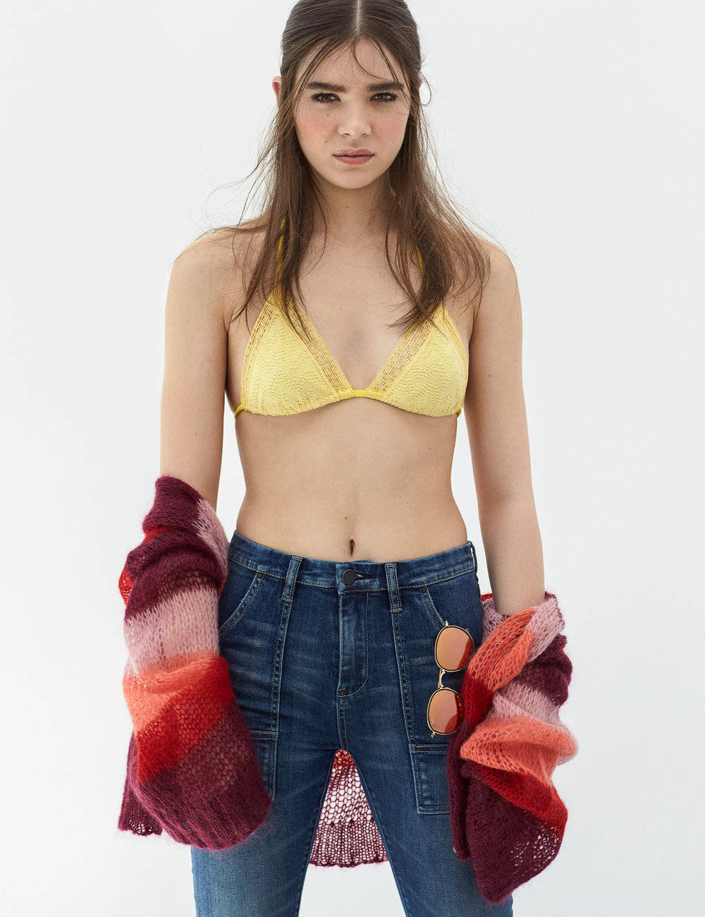 Hailee Steinfeld《Flaunt Magazine》封面 壁紙 — 一位穿著黃色蕾絲比基尼上衣和牛仔褲的女性，在純白背景前，手持一條色彩繽紛的針織披肩，口袋裡放著太陽眼鏡。