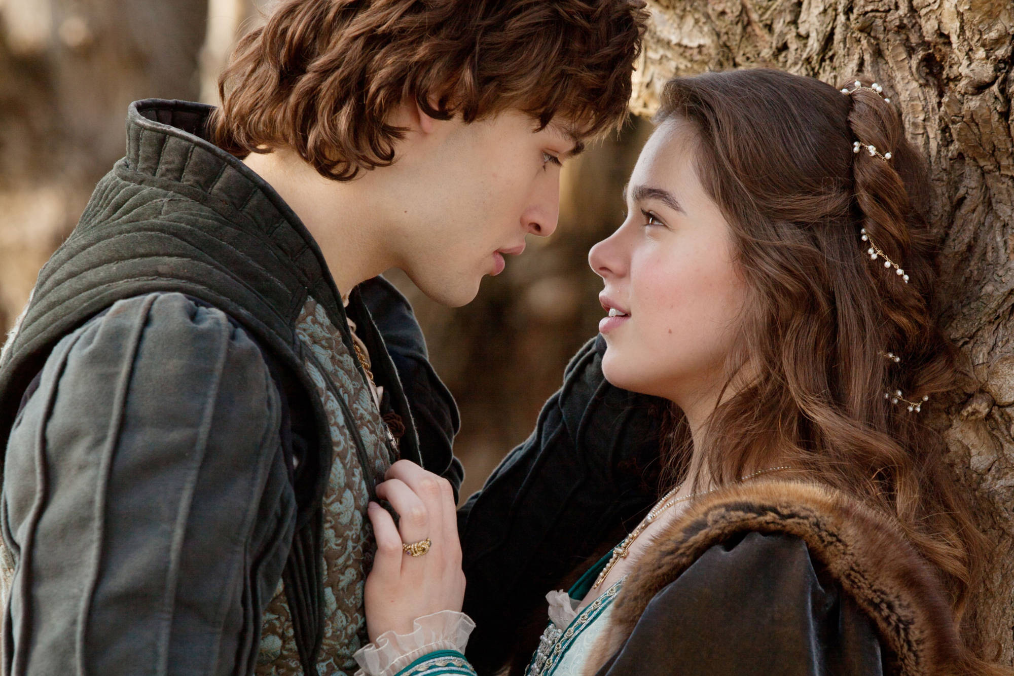 Hailee Steinfeld與Douglas Booth《羅密歐與茱麗葉》 壁紙 — 一對穿著古裝的年輕情侶在陽光普照的森林空地樹下分享親密時刻。