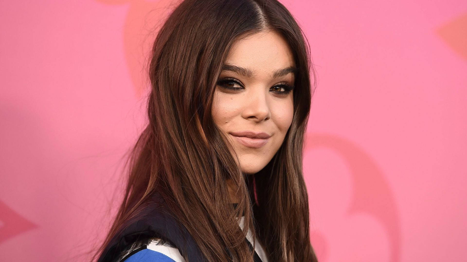 Hailee Steinfeld路易威登活動 壁紙 — 一位長髮棕色的年輕女性在帶有微妙心形圖案的鮮豔粉紅色背景前溫柔微笑。