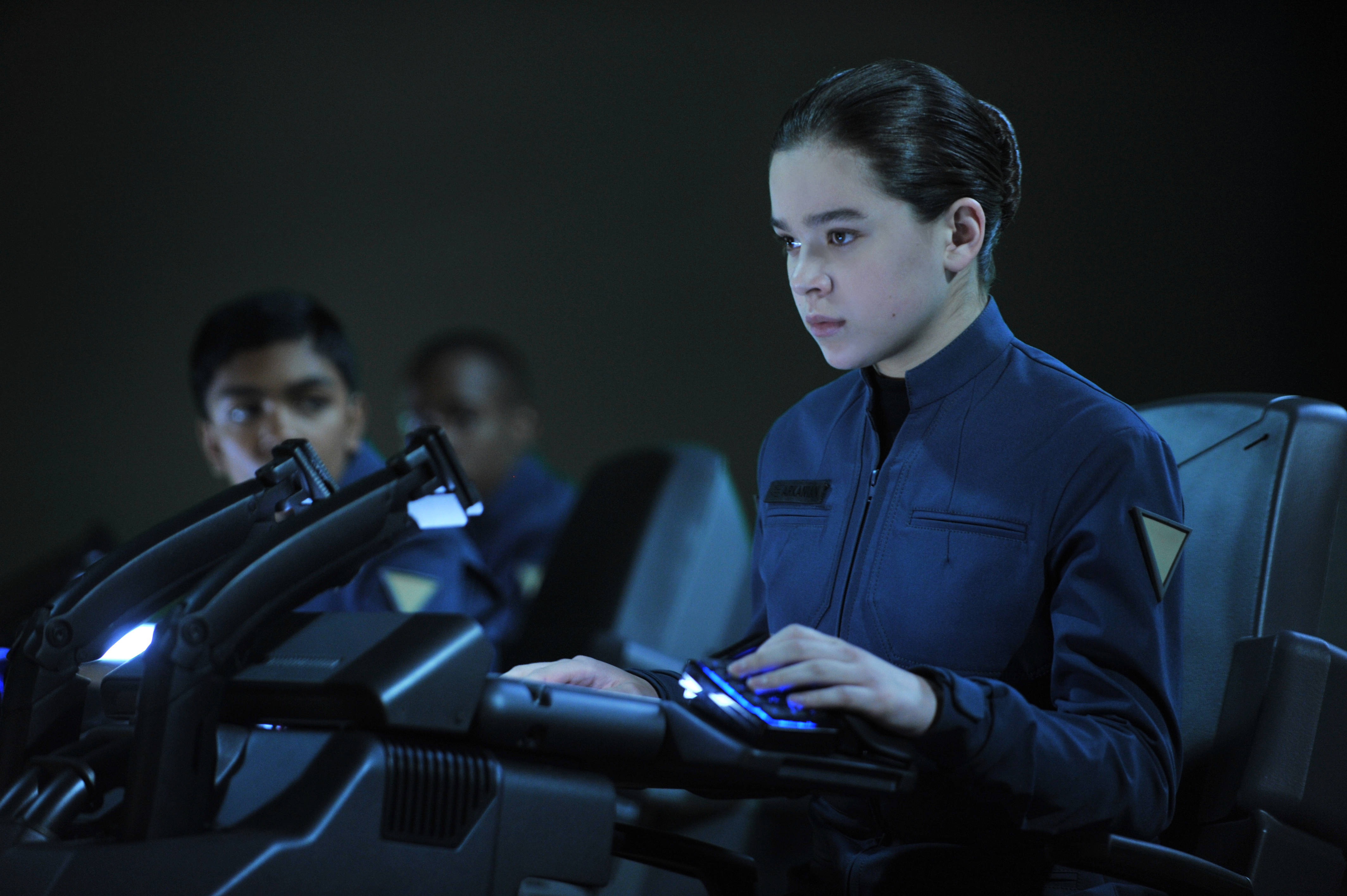Hailee Steinfeld《Ender's Game》彼得拉·阿爾卡尼安 壁紙 — 一位身穿深藍色制服嘅年輕太空人，喺未來感嘅駕駛艙中，專注於發光嘅控制面板，燈光昏暗帶藍色調。