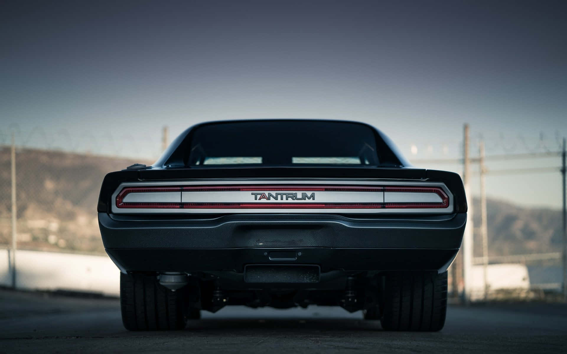  壁紙 — 黑色Dodge Challenger Tantrum停泊在賽道上，背景有鐵絲網圍欄和山丘的後視圖。