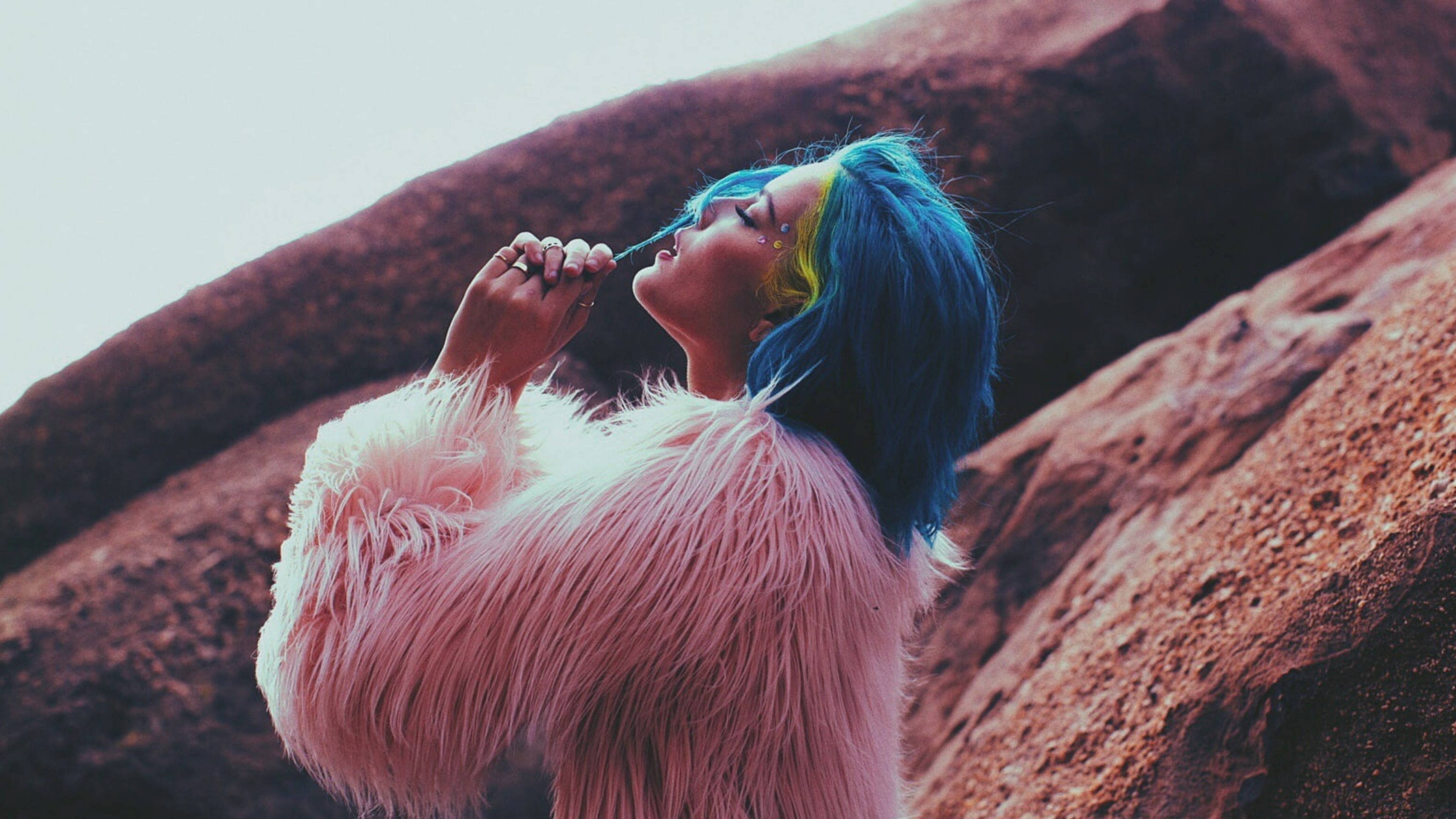Halsey《Badlands》專輯造型 壁紙 — 一個藍黃色頭髮的人穿著毛茸茸的粉紅色外套，在微紅色的岩層背景下吸煙。