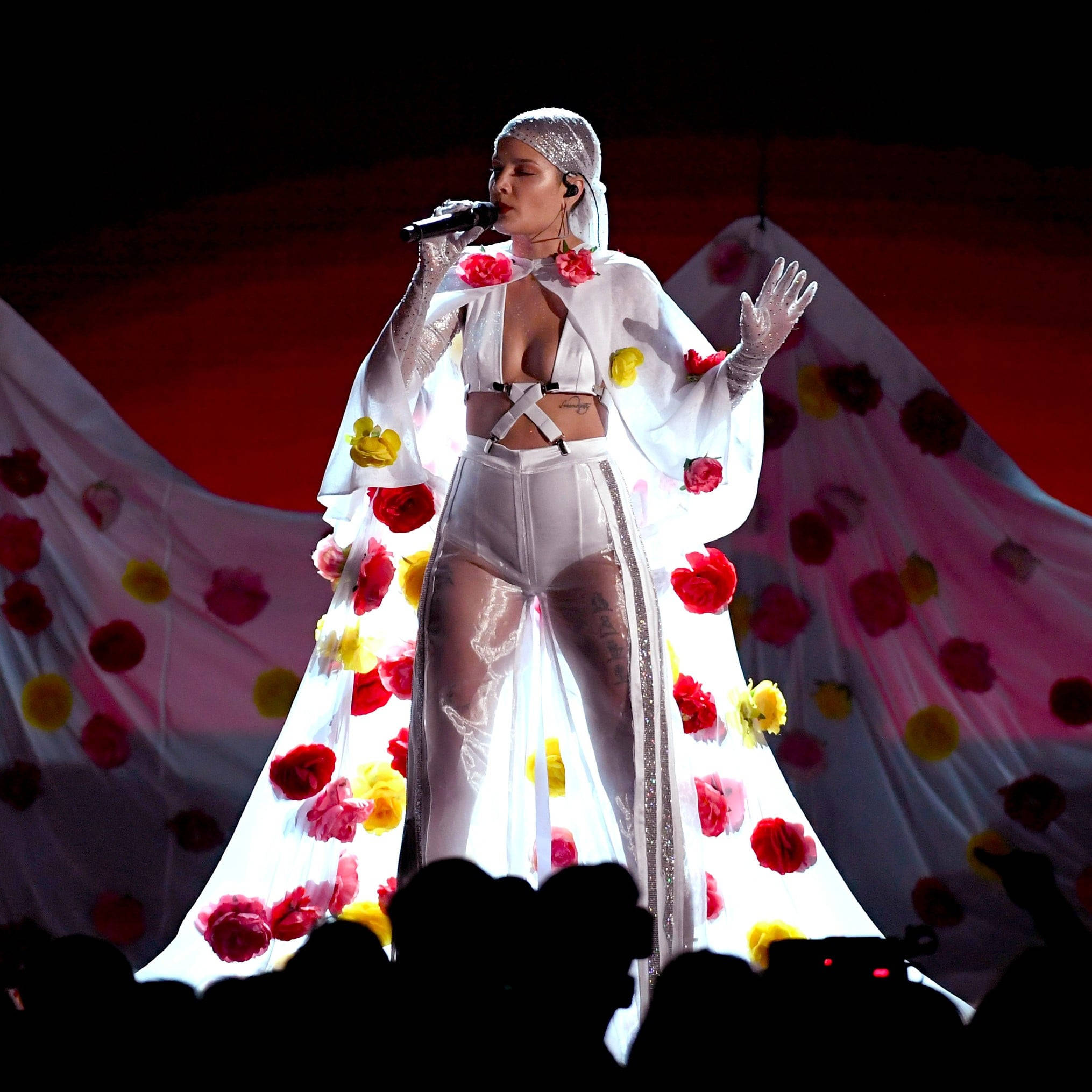 Halsey 在 2017 年 Billboard 音樂獎驚豔亮相 壁紙 — 一位身穿白色花卉長裙的歌手在明亮的燈光下於舞台上表演，背景為紅色，下方有觀眾剪影。