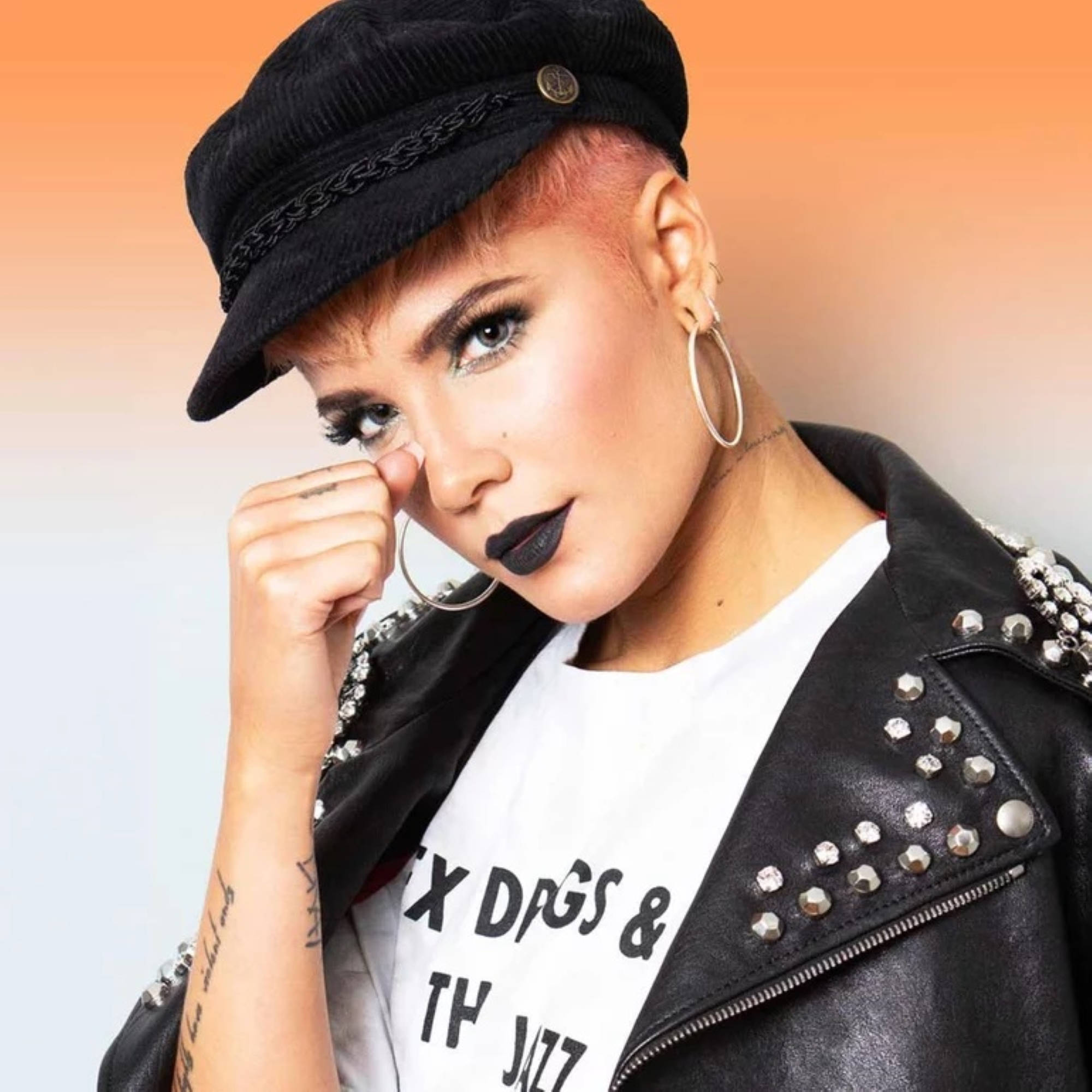 Halsey 妝容黑唇 壁紙 — 一位大膽的女性，粉紅色頭髮，戴黑色帽子，穿著鉚釘皮外套，在溫暖的漸變背景前自信地擺姿勢。