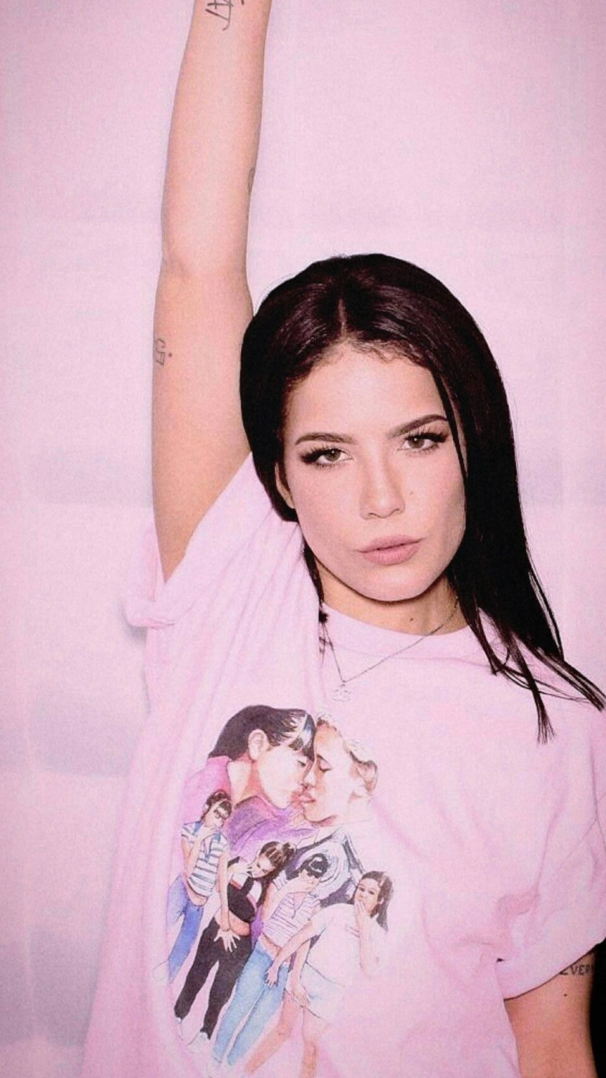Halsey 的休閒造型 壁紙 — 一位深色頭髮、有紋身的年輕女性，穿著圖案T恤，在柔和的粉紅色背景前舉起手臂。