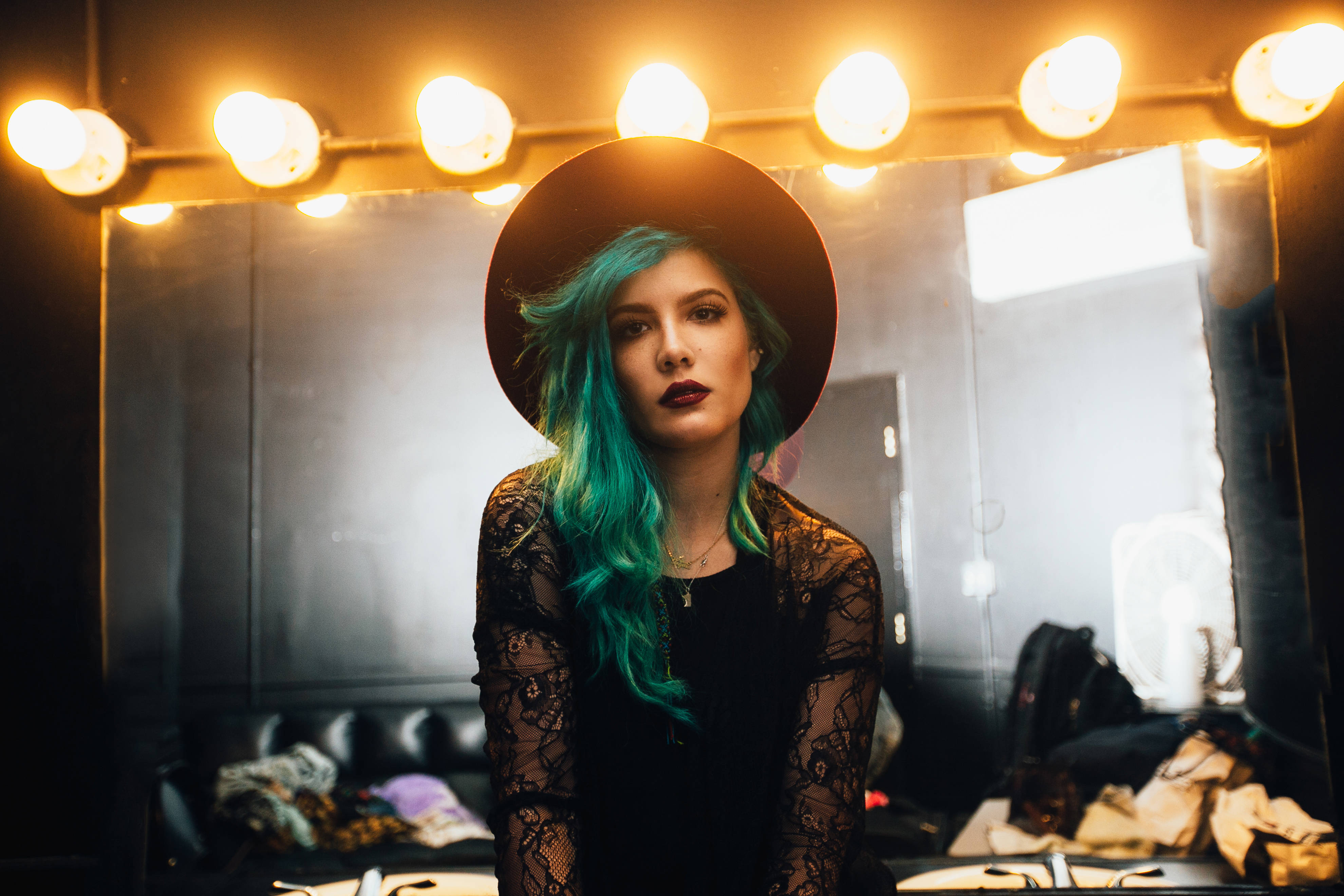 Halsey 穿黑色蕾絲禮服 壁紙 — 一位擁有充滿活力的藍綠色頭髮和寬邊帽的女性，在後台化妝間溫暖的化妝燈下直視鏡頭。