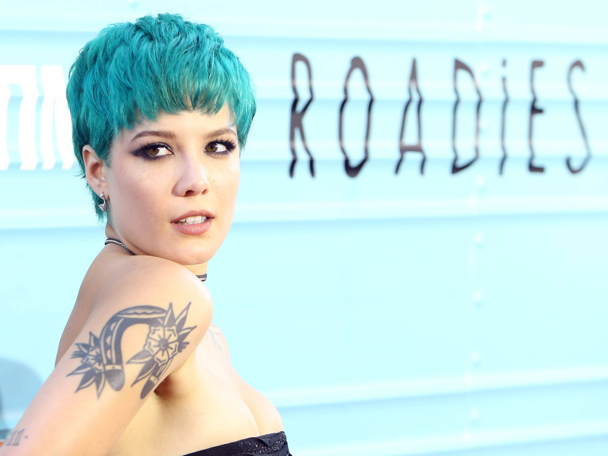 Halsey 青綠髮色 壁紙 — 一位擁有充滿活力的藍綠色短髮和詳細肩部紋身的女性，在淺藍色 'ROADIES' 背景前擺姿勢。