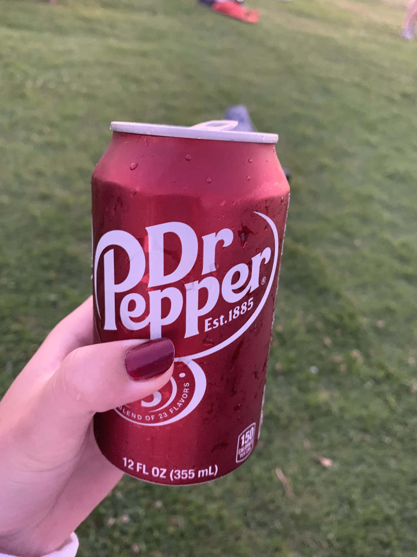  壁紙 — 一隻塗有栗色指甲油的手，拿著一罐冰凍的Dr Pepper，罐身凝結著水珠，背景是模糊的綠色草地。