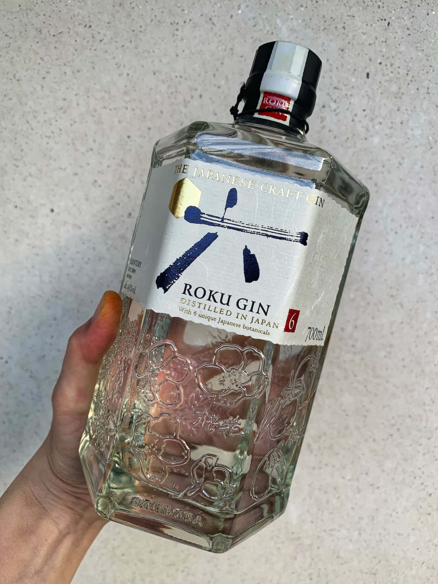  壁紙 — 一隻手拿著一個透明的六角形Roku Gin酒瓶，背景為灰色斑點，展示其日本工藝標籤和花卉蝕刻。