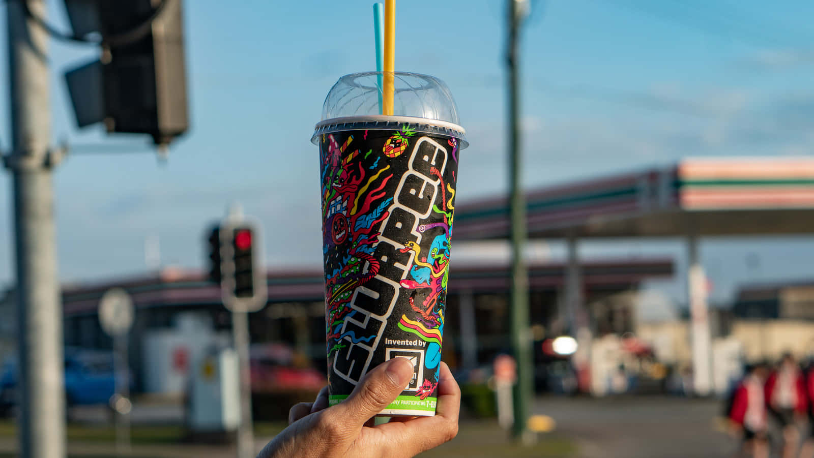  壁紙 — 一隻手拿著一個充滿活力的塗鴉風格 Slurpee 杯，配有兩根吸管，背景是模糊的加油站，天空清澈湛藍。