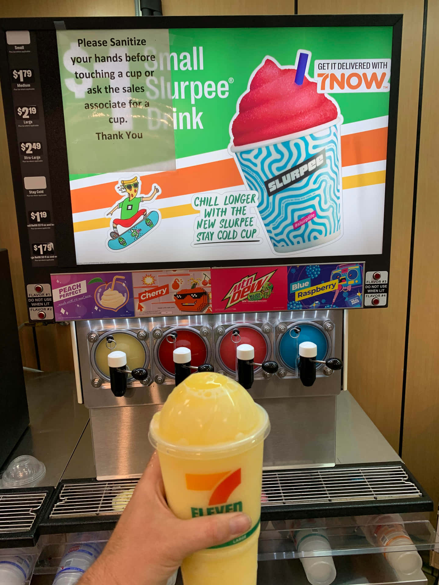  壁紙 — 一隻手在7-Eleven思樂冰機前拿著黃色思樂冰，明亮的數碼廣告顯示口味和價格。