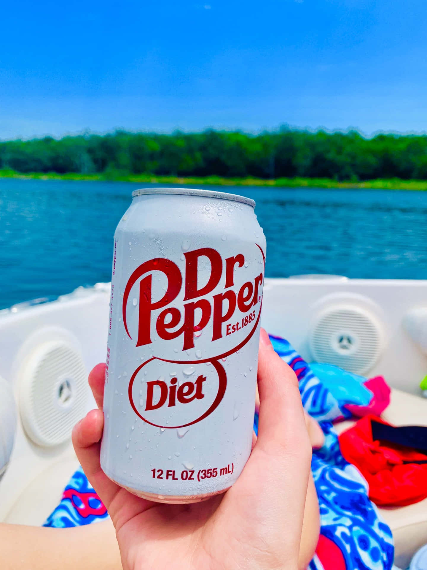  壁紙 — 一隻手喺船上拎住一罐冰凍嘅Diet Dr Pepper，背景係藍色嘅水同綠色嘅樹，天空晴朗。