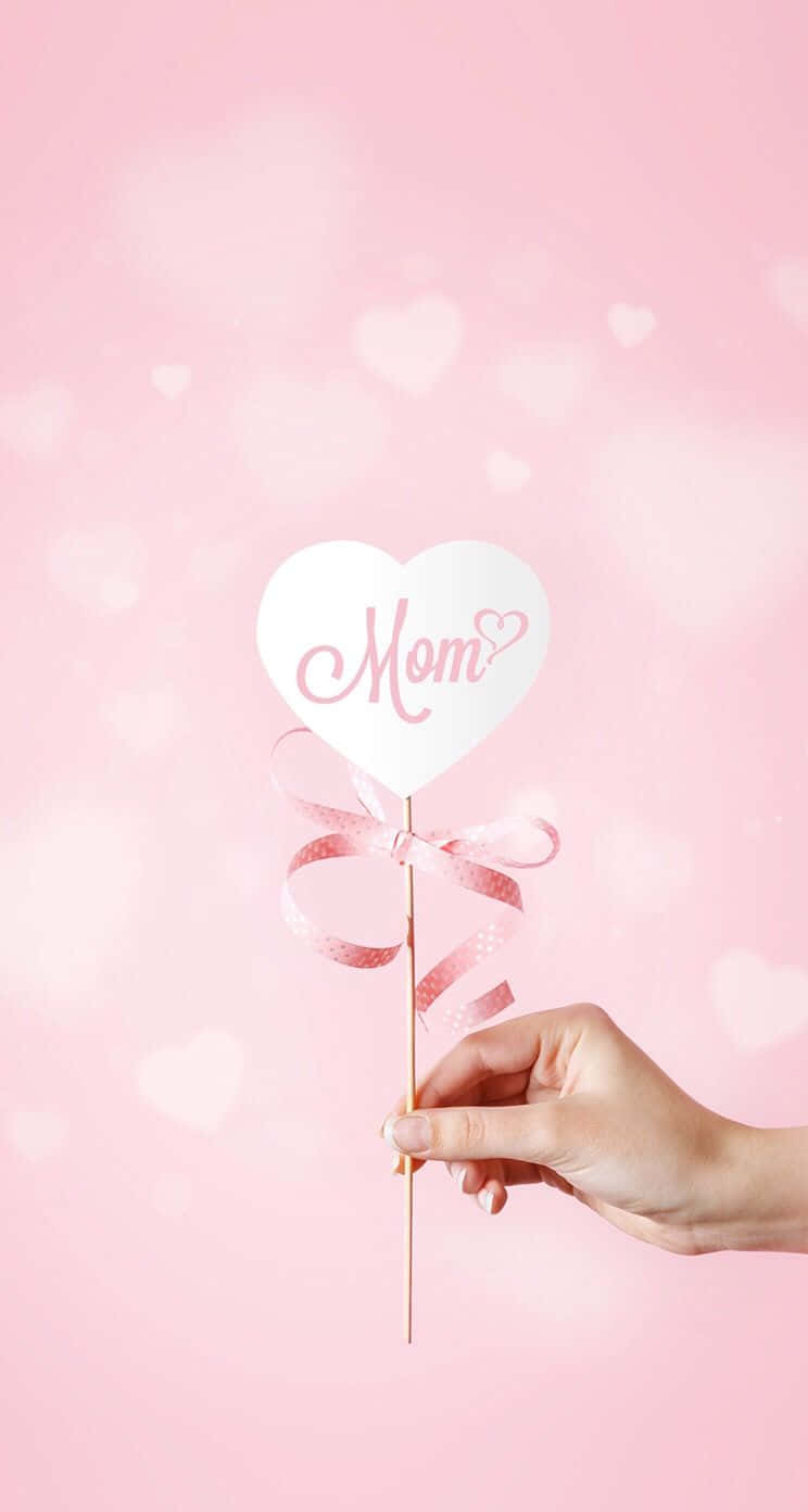  壁紙 — 一隻手拿著一個白色心形標牌，上面用粉紅色草書寫著「Mom」，並繫著粉紅色絲帶，背景是柔和的粉紅色，帶有模糊的心形散景。