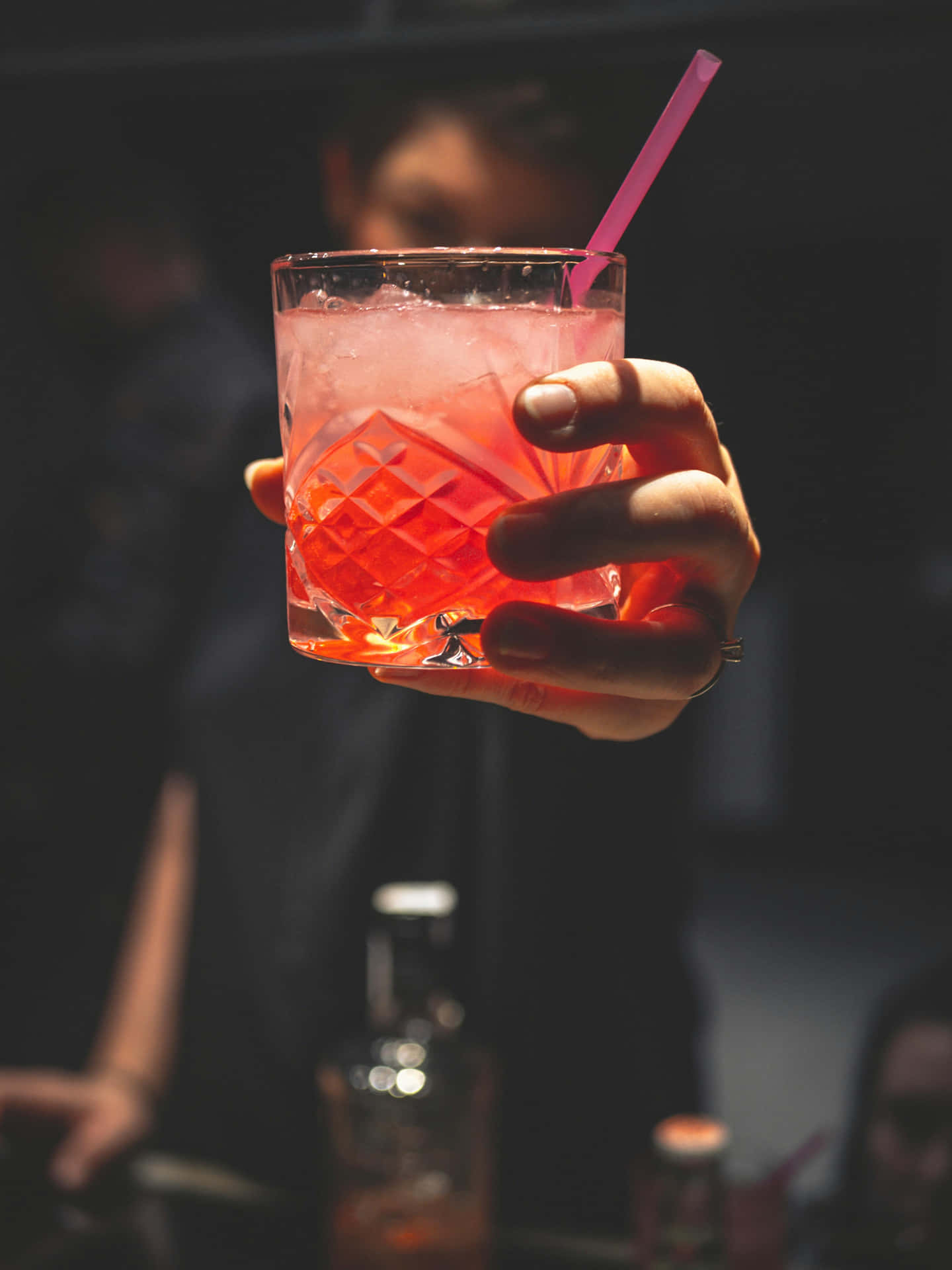  壁紙 — 一隻手喺昏暗嘅酒吧背景下，用一個有紋理嘅玻璃杯，攞住一杯充滿活力嘅粉紅色雞尾酒，仲有粉紅色飲管。