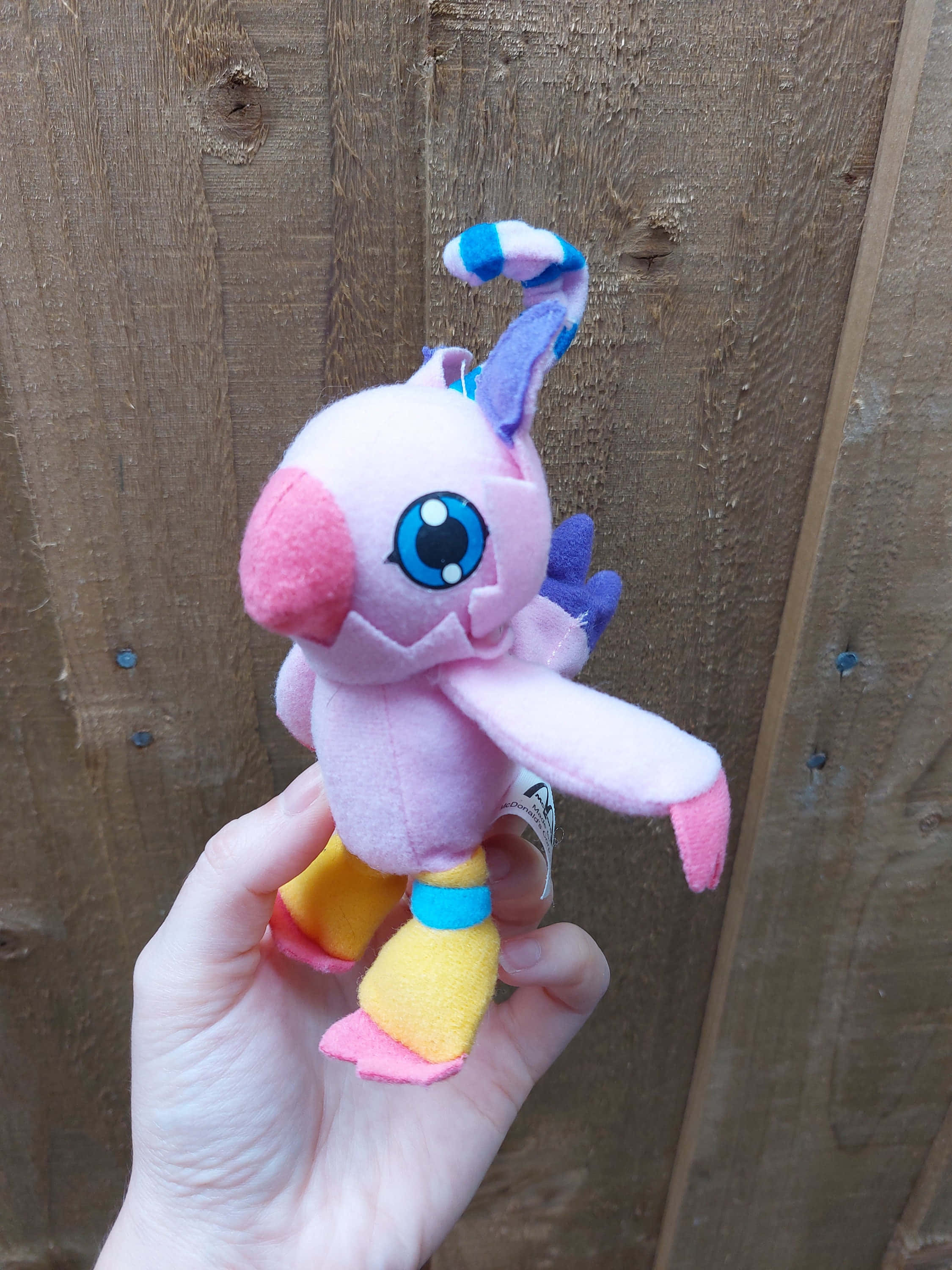 Digimon Biyomon 桌布 壁紙 — 一隻手拎住一個粉紅色毛絨玩具，有藍色眼睛、紫色耳朵同黃色腳，背景係木欄柵。