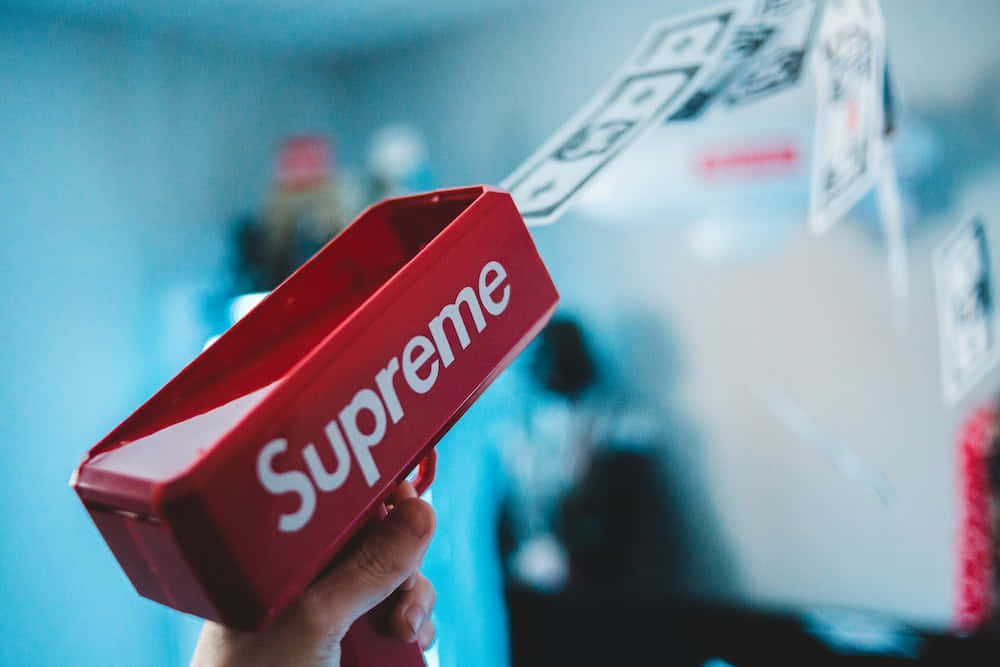 手拿著紅色Supreme玩具槍，美元鈔票在藍色背景前飛舞。
