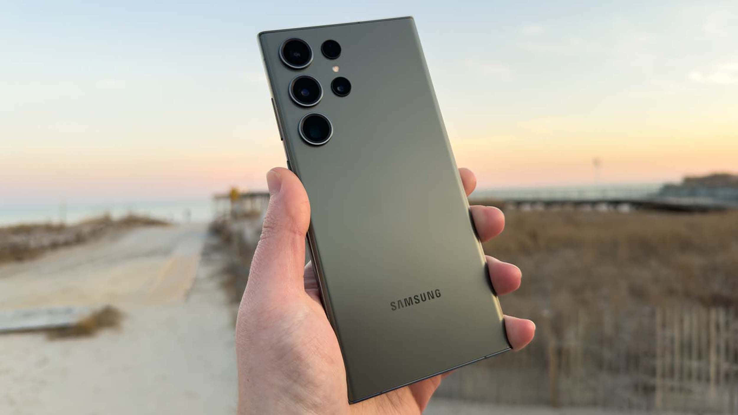 Samsung S23 Ultra 桌布 壁紙 — 一隻手拿著一款時尚的橄欖綠色Samsung智能手機，背景是柔和的海灘日落，帶有模糊的沙丘和海洋地平線。