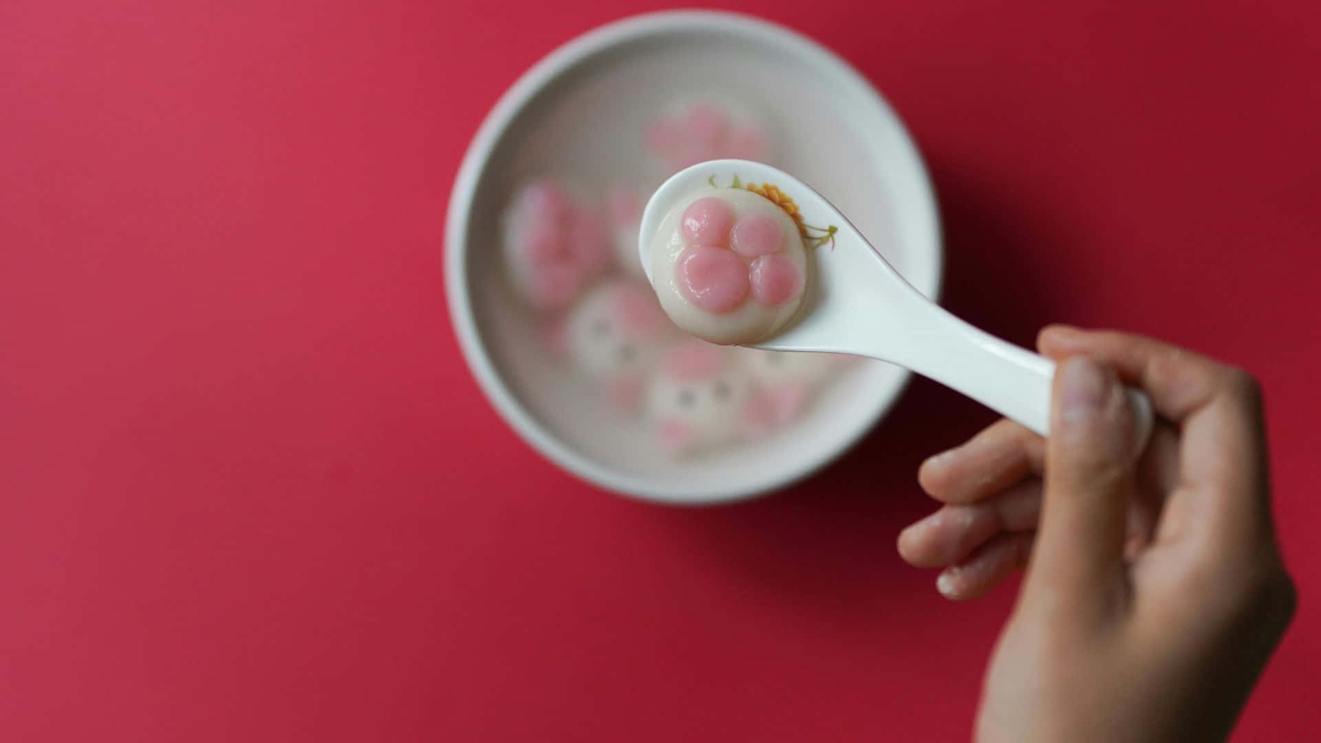  壁紙 — 一隻手拿著湯匙，從白色碗中舀起一個粉紅色貓臉餃子，背景為鮮紅色。