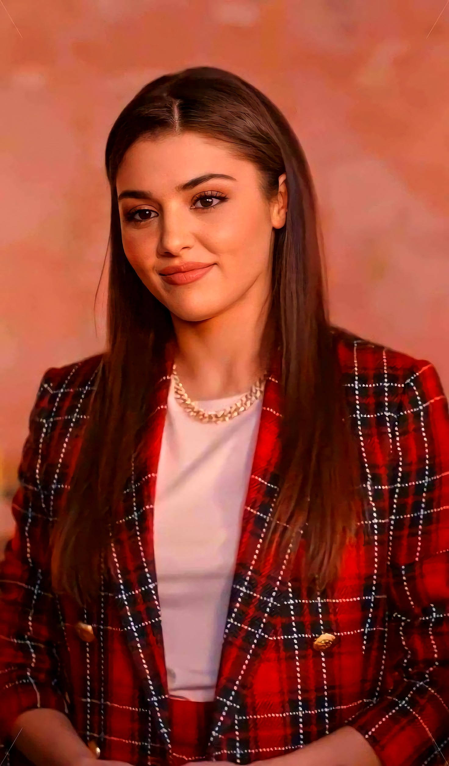 Hande Ercel 微笑 壁紙 — 一位長棕髮女性在溫暖的桃色背景前，身穿紅色格紋西裝外套，內搭白色上衣。
