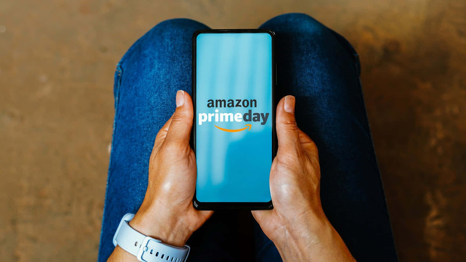  壁紙 — 雙手握著智能手機，顯示Amazon Prime Day標誌，置於穿著牛仔褲的膝蓋上，背景溫暖有質感。