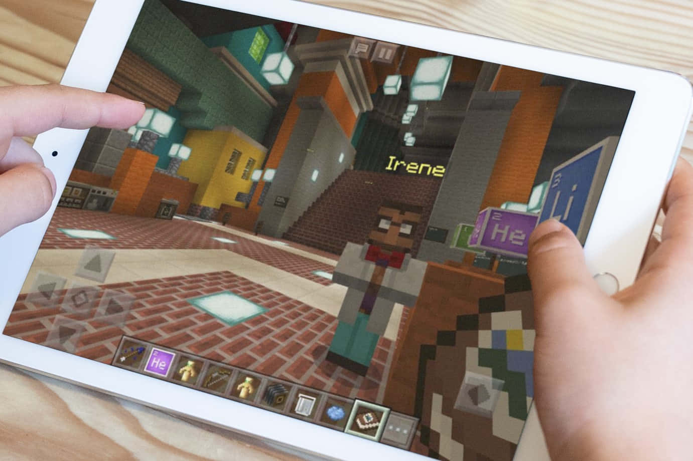 Minecraft Education Edition 桌布 壁紙 — 雙手拿著平板電腦，顯示《Minecraft》遊戲場景，有一個名為 Irene 的像素角色在方塊狀的室內環境中。