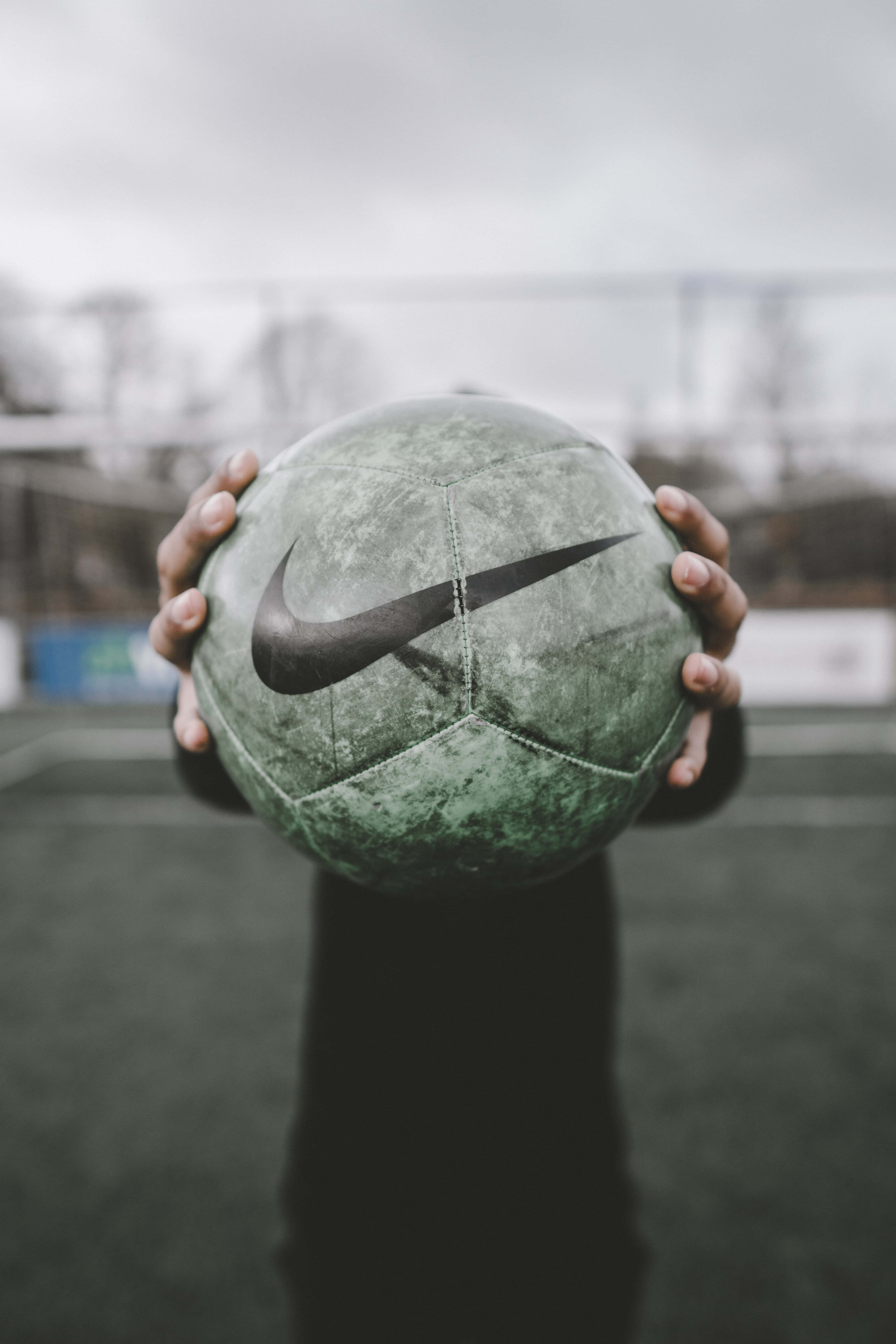 ball 桌布 壁紙 — 雙手在陰天嘅戶外球場上，拎住一個風化咗嘅綠色Nike足球，背景模糊。