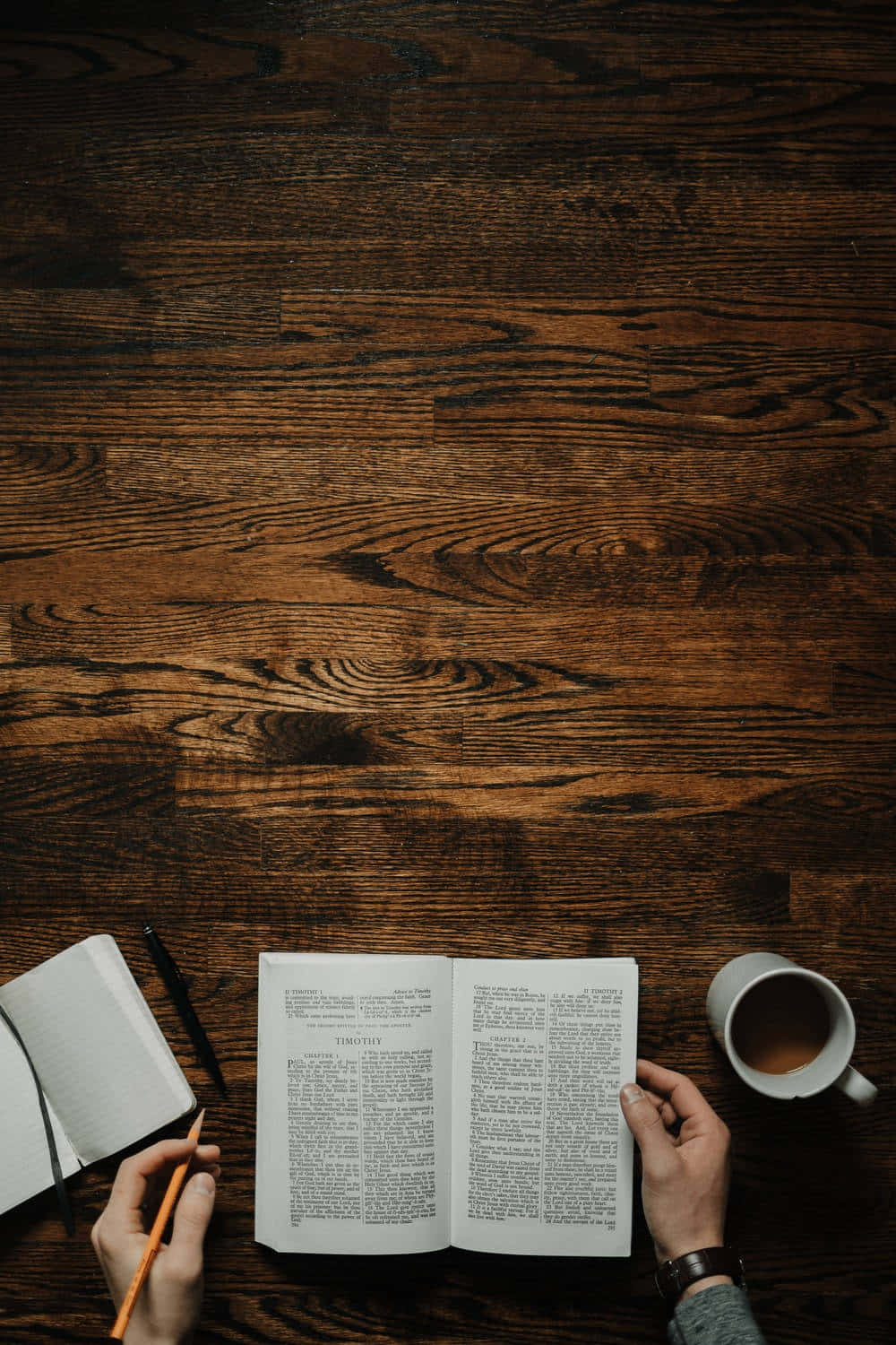 Dark Book Aesthetic 桌布 壁紙 — 雙手在深色木桌上，咖啡杯旁，拿著一本打開的書和鉛筆。