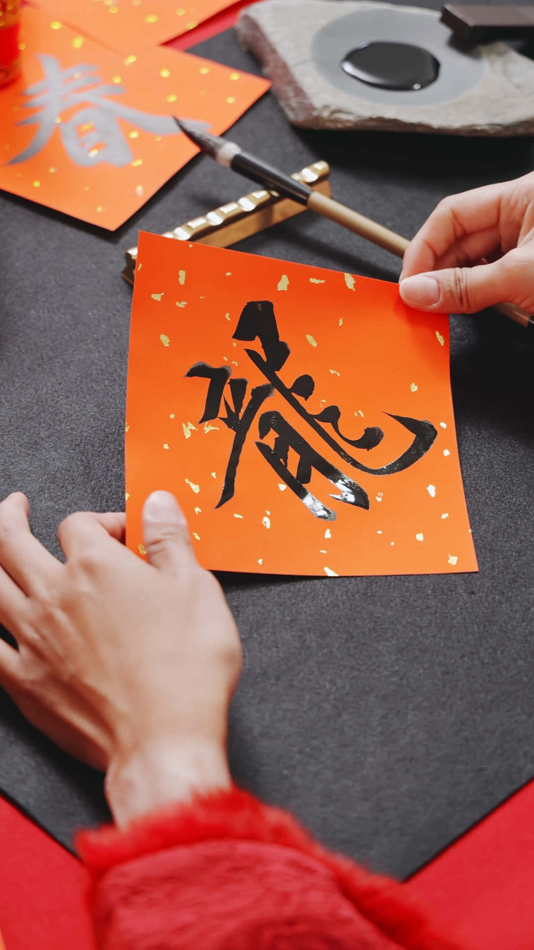  壁紙 — 雙手小心地拿著一張橙色紙，上面有粗體黑色中國書法，背景是深色紋理表面。