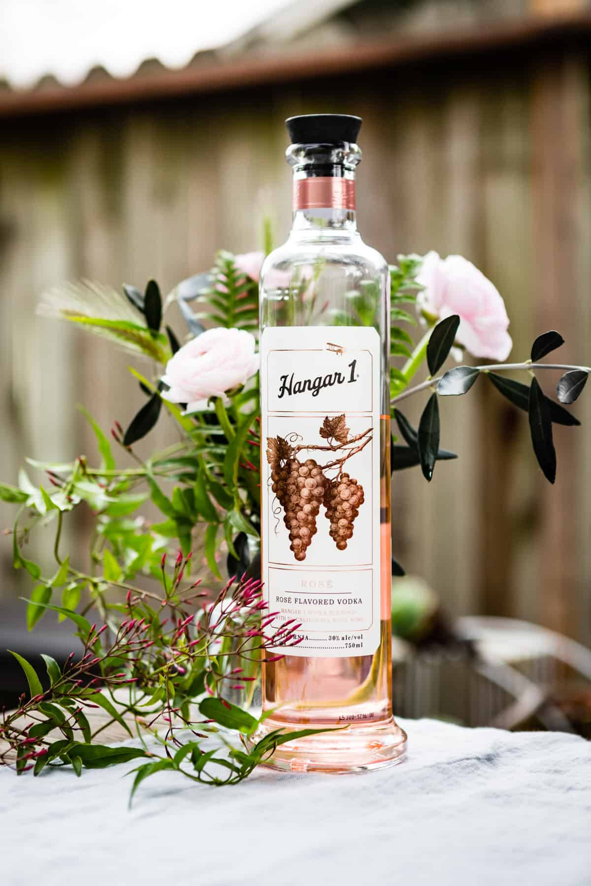 Hangar 1 銀玫瑰金葡萄柚花 壁紙 — 一瓶 Hangar 1 Rose Flavored Vodka 優雅地放在大理石表面上，周圍環繞著柔和的粉紅色花朵和綠色植物。