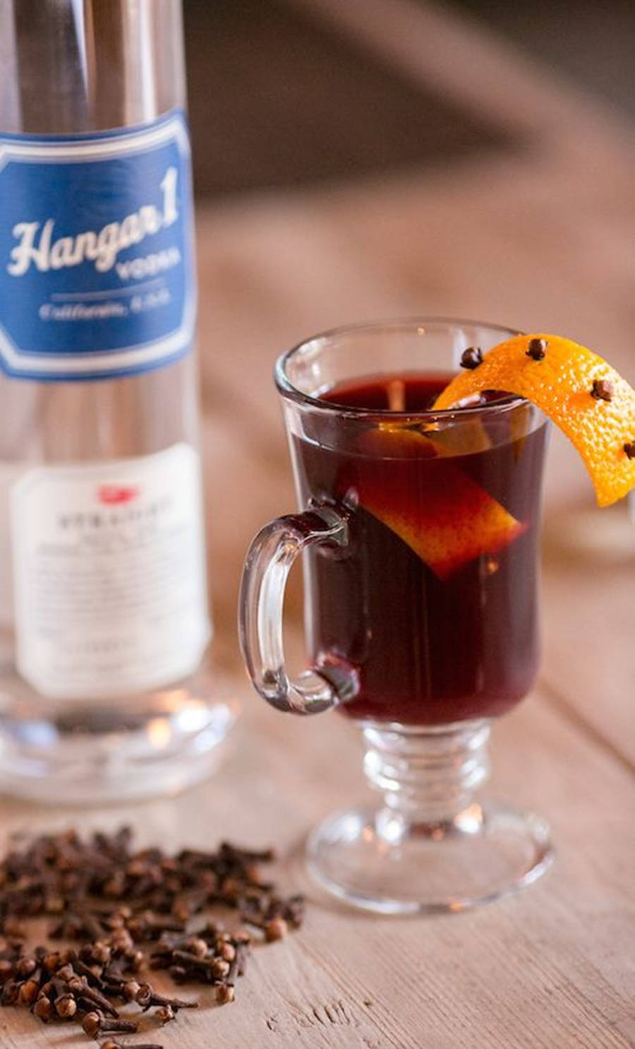 Hangar 1三款飲品 壁紙 — 一杯加了香料嘅深紅色熱紅酒，用橙片同丁香裝飾，擺喺木枱上嘅Hangar 1伏特加酒樽旁邊。