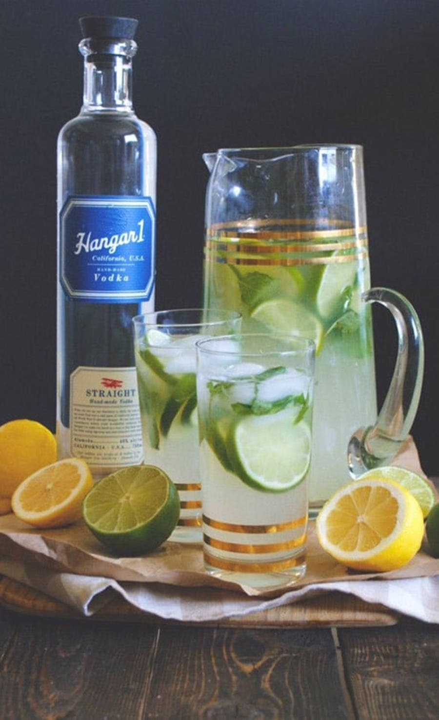 Hangar 1頂級美國伏特加優雅倒酒 壁紙 — 一個清爽的青檸和檸檬伏特加飲品壺，配有冰塊和柑橘片，放在質樸的木桌上。