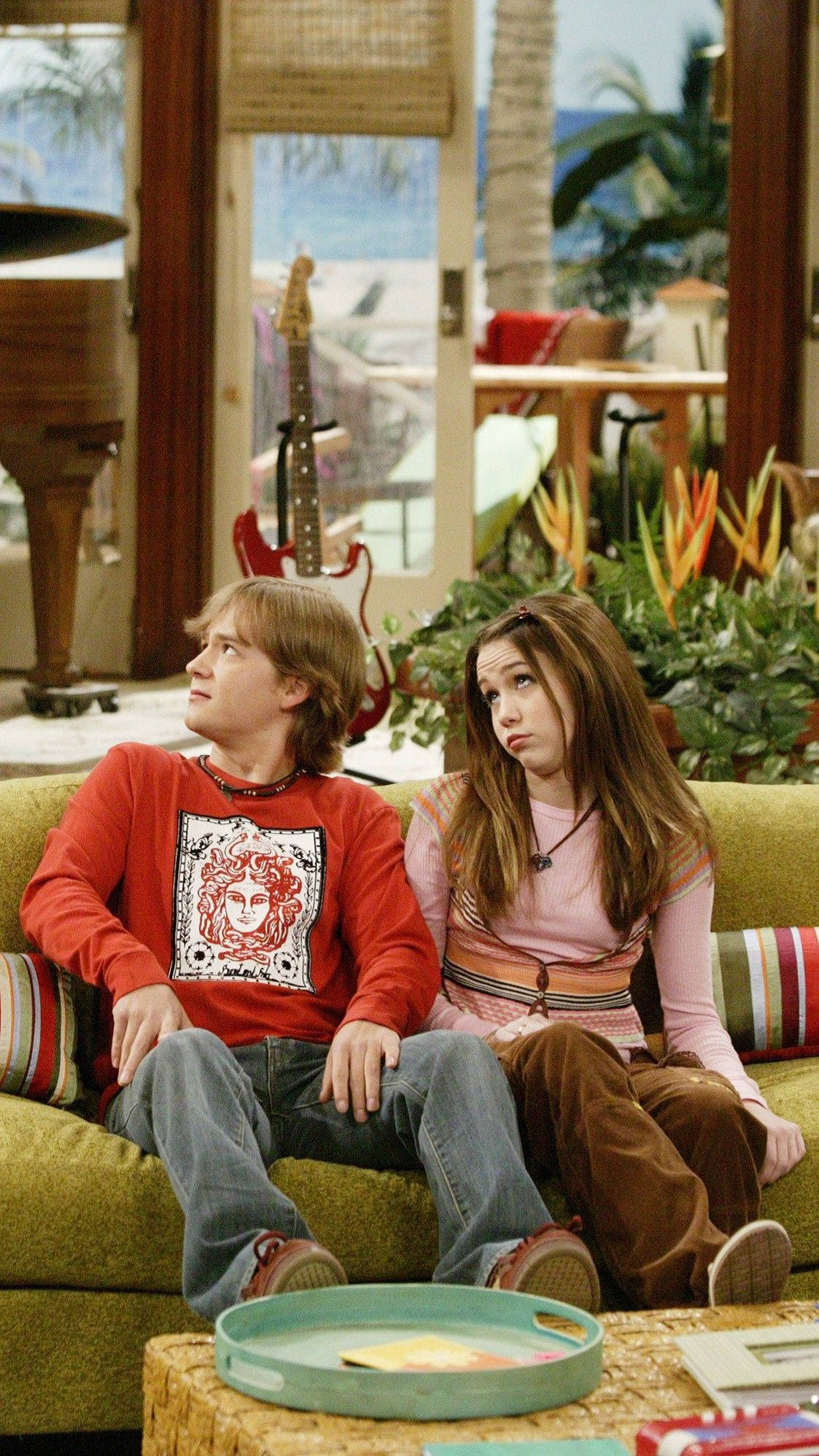 Hannah Montana 搞笑表情 壁紙 — 兩個青少年坐在一個熱帶主題客廳的綠色沙發上，可以看到棕櫚樹和結他。