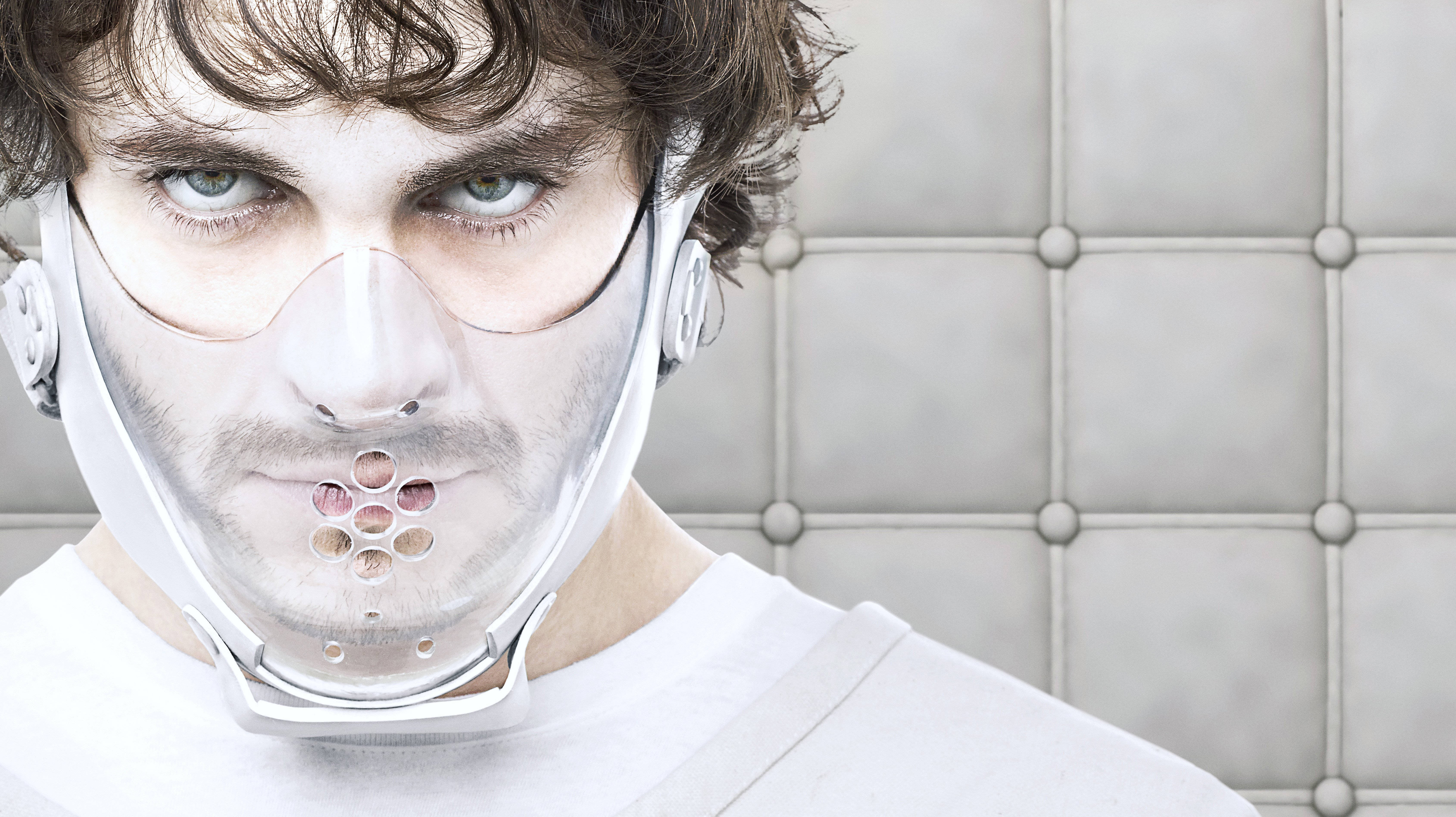 《 Hannibal 》威爾戴著嘴套 壁紙 — 一名眼神銳利的男子戴著未來風格的透明面罩，上面有圓形通風口，背景是簇絨灰色牆壁。