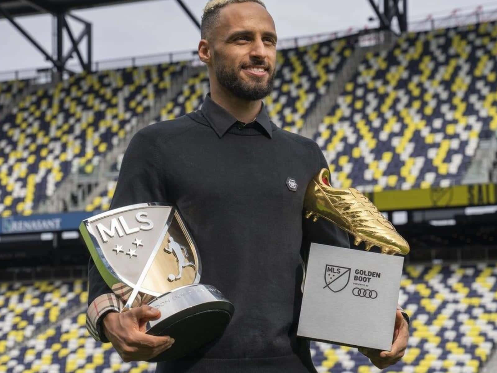 Hany Mukhtar 拿著2022年Landon Donovan MLS MVP獎盃 壁紙 — 一位微笑的足球運動員在一個空蕩蕩的體育館內自豪地高舉兩個MLS獎盃，座位上黃藍相間。