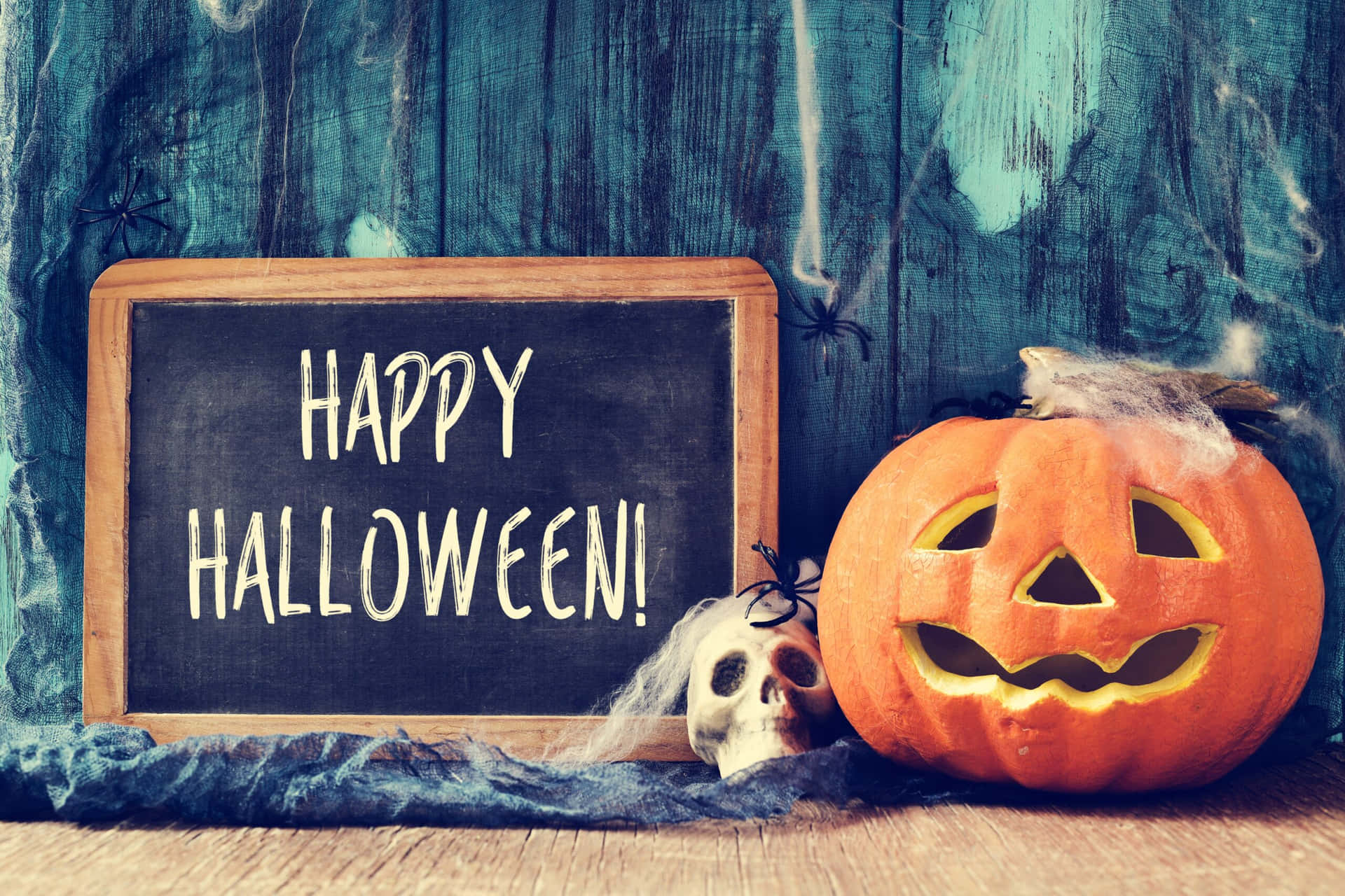  壁紙 — 一個雕刻的橙色南瓜燈，帶有發光的眼睛，旁邊放著一塊寫有「HAPPY HALLOWEEN!」的黑板，還有一個被蜘蛛網覆蓋的頭骨，背景是風化的藍色木牆。
