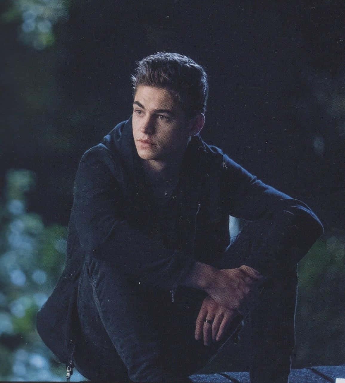 Hardin Scott 的特寫：陰影中的孤獨 壁紙 — 一個穿著深色連帽衫的年輕人在夜晚戶外沉思地坐著，身後有模糊的綠色植物。