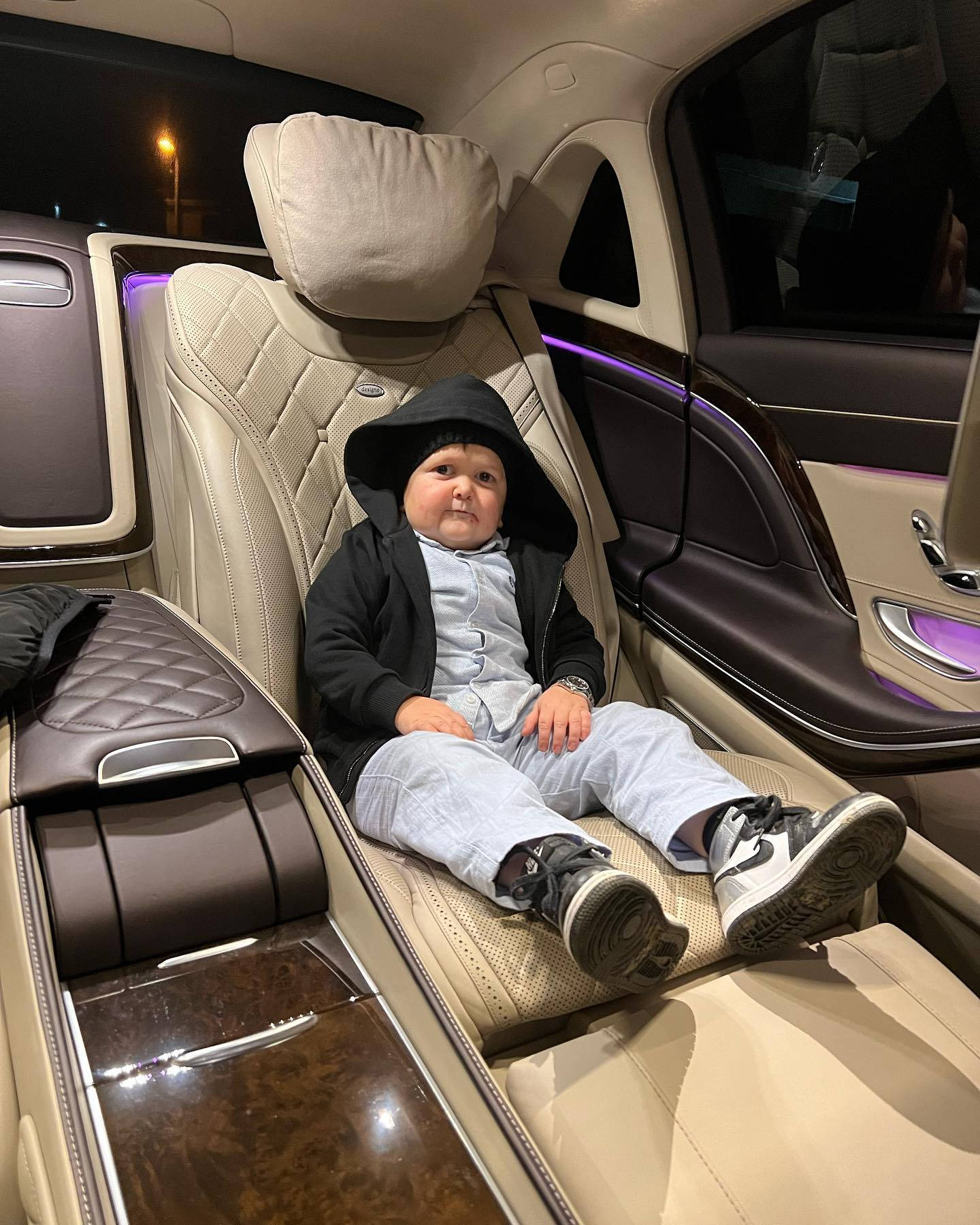 Hasbulla 坐在豪華車後座 壁紙 — 一個穿著黑色連帽外套的幼兒自信地坐在一輛高級汽車的豪華後座，車內有紫色氛圍燈。