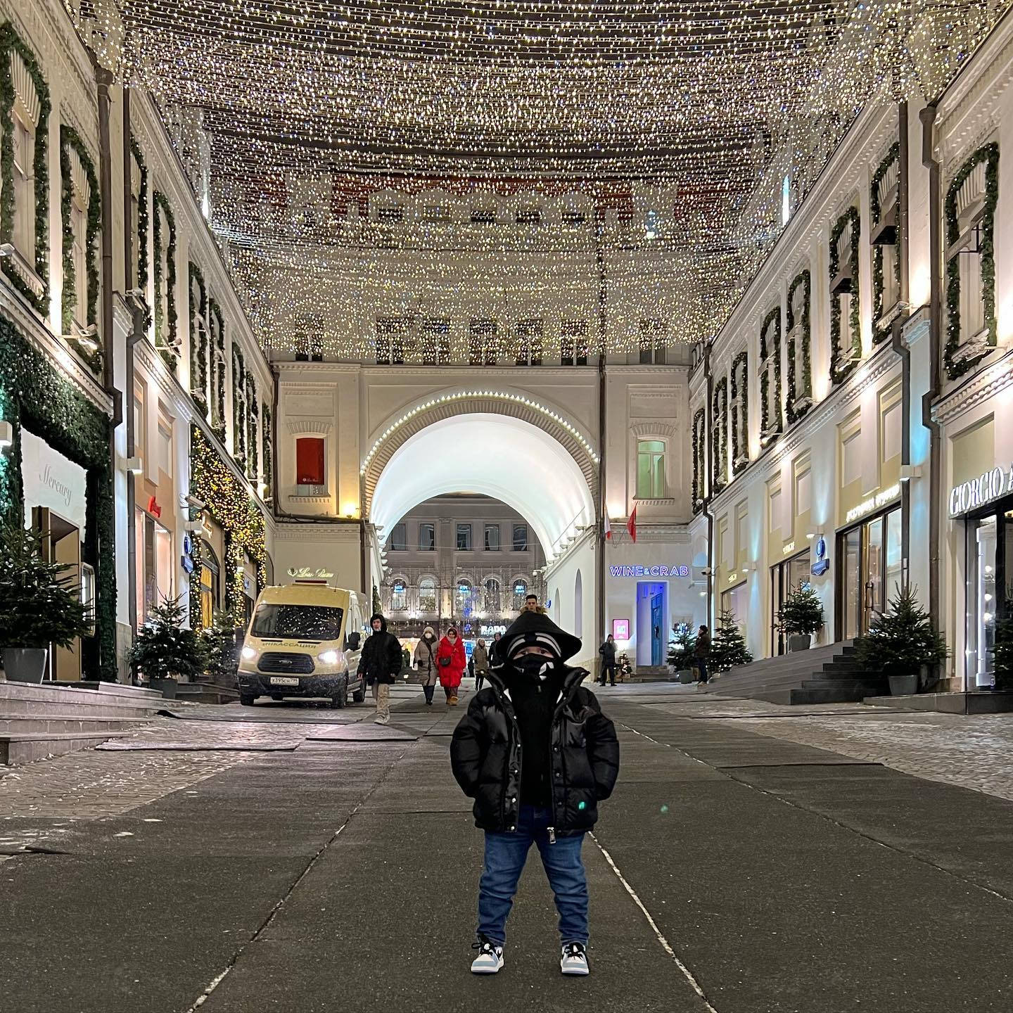 Hasbulla 穿著冬季服裝 壁紙 — 一個穿著黑色羽絨外套的孩子，在夜晚宏偉的城市拱廊下，站在耀眼的聖誕燈飾天篷中央。