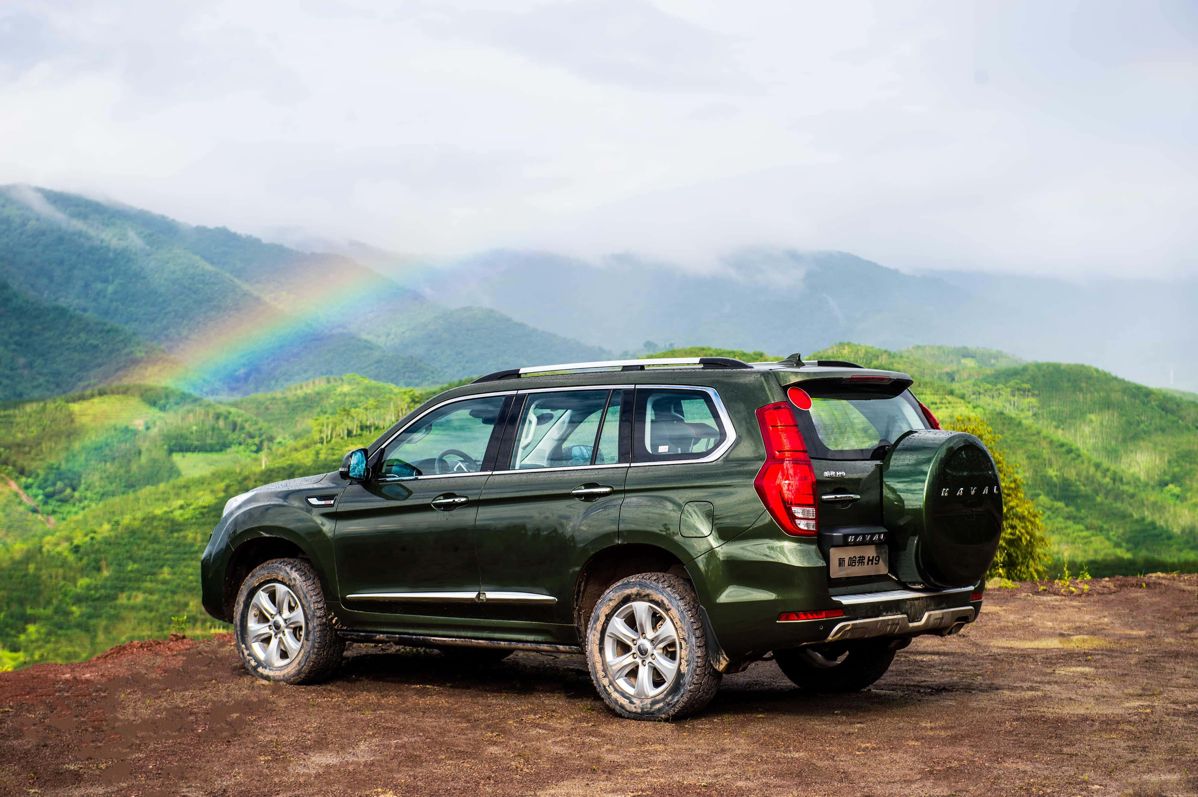 時尚強悍的 Haval SUV 在路上 壁紙 — 一架深綠色SUV停泊在泥濘的觀景點，彩虹在雲天之下拱起於翠綠山巒之上。